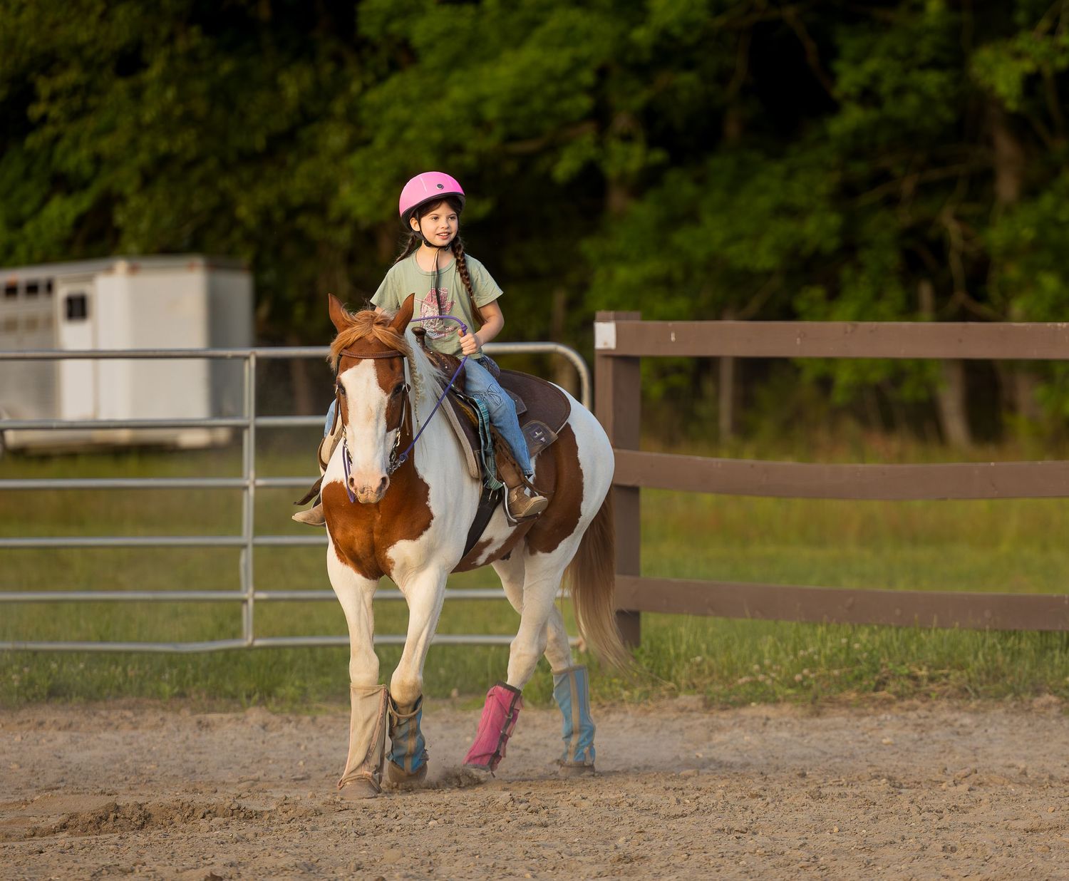 Young girl in pink helmet rides a brown and white paint horse in a sandy riding arena with green trees in background.