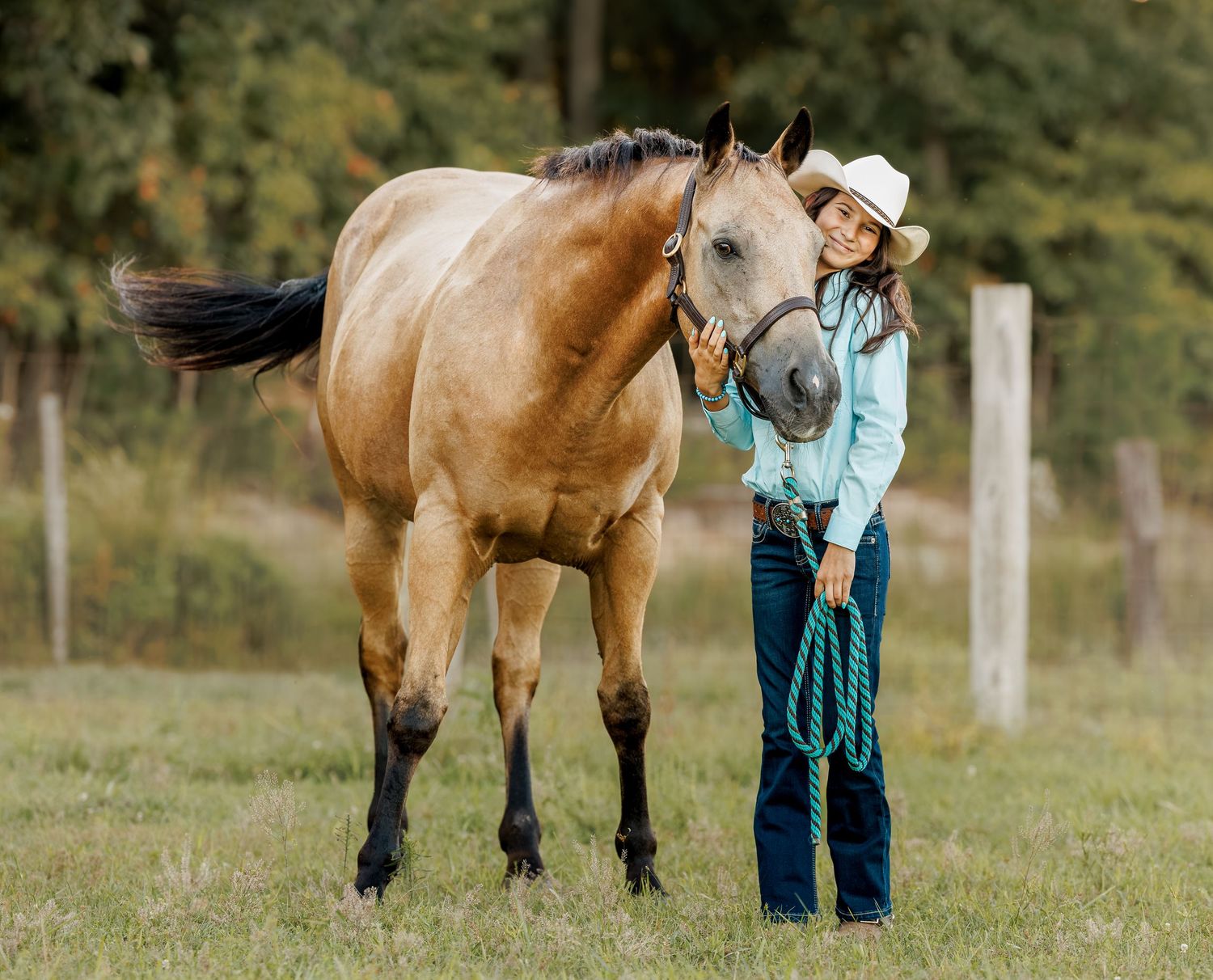 Young woman in cowboy hat and turquoise shirt stands beside a buckskin horse on a green pasture.