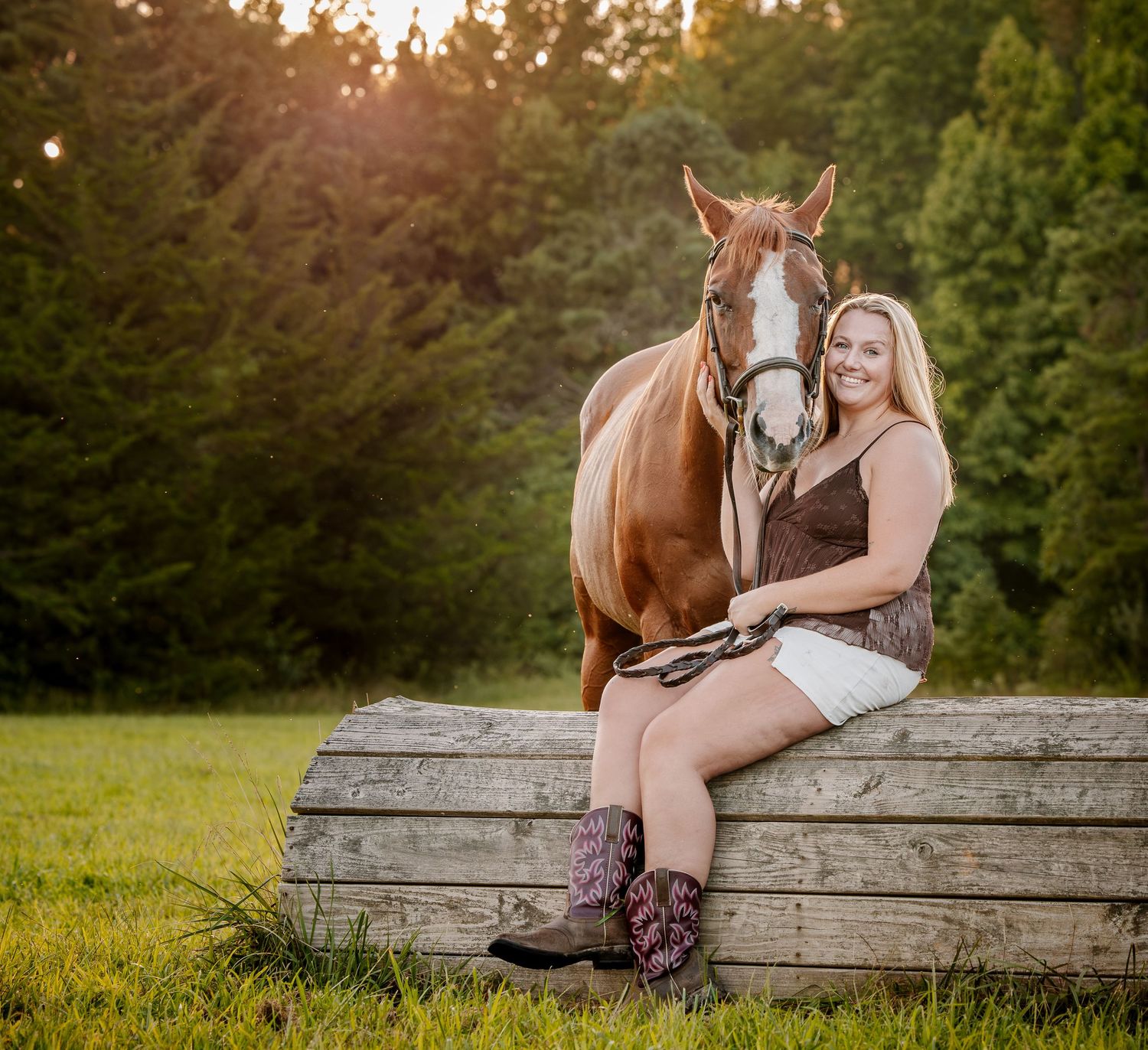 Young woman in cowboy boots and white shorts sits smiling beside a chestnut horse on a wooden fence at golden hour.