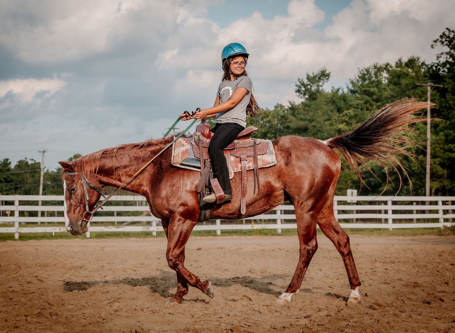 Young girl in blue helmet riding a chestnut horse in a fenced arena on a sunny day.