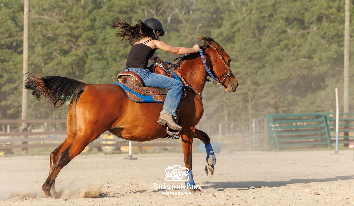 A young woman in a black helmet rides a bay horse during a barrel racing event in a sandy arena.