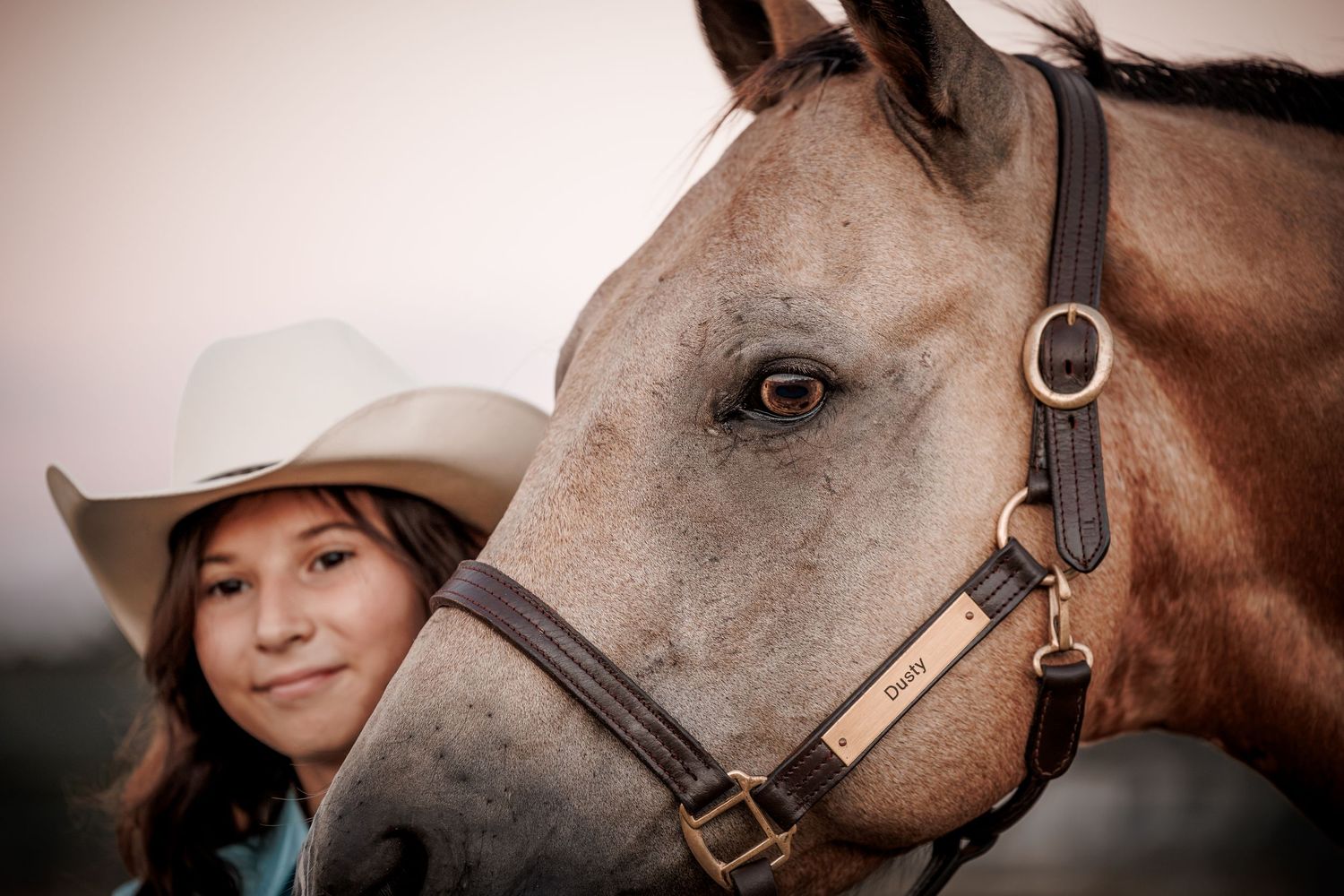Young woman in white cowboy hat poses closely with a gray horse wearing a leather halter outdoors.