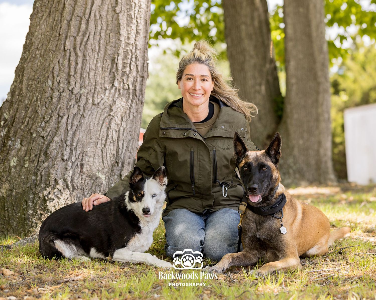 Woman smiling outdoors with a Border Collie and Belgian Malinois beside large trees, professional dog photography session.