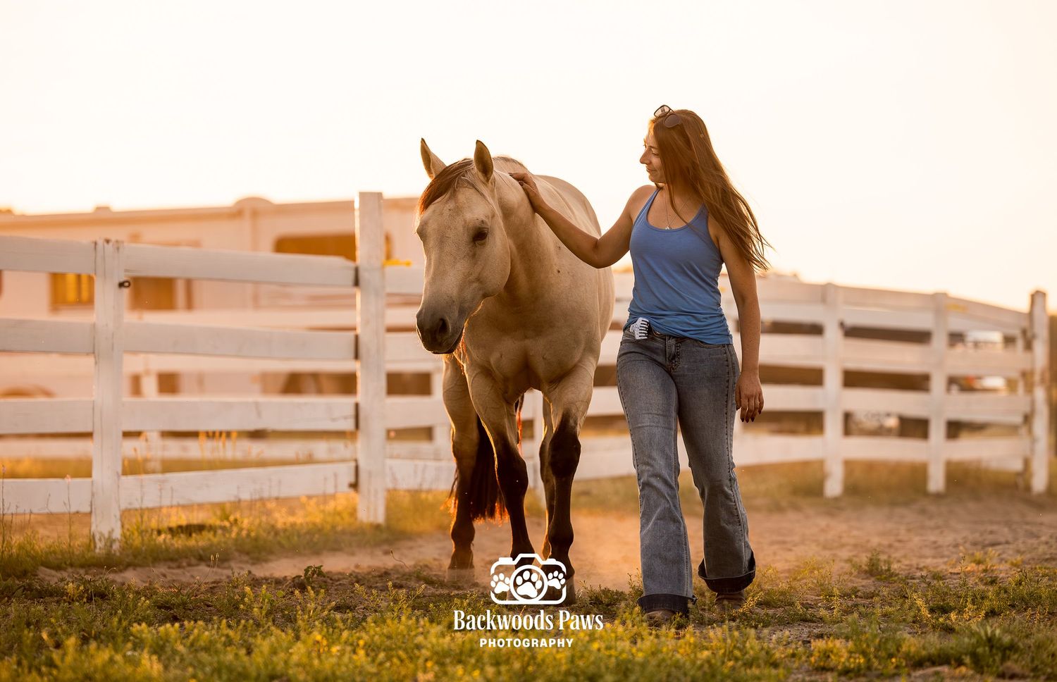 Woman walking alongside a palomino horse in a white-fenced paddock at golden hour sunset.