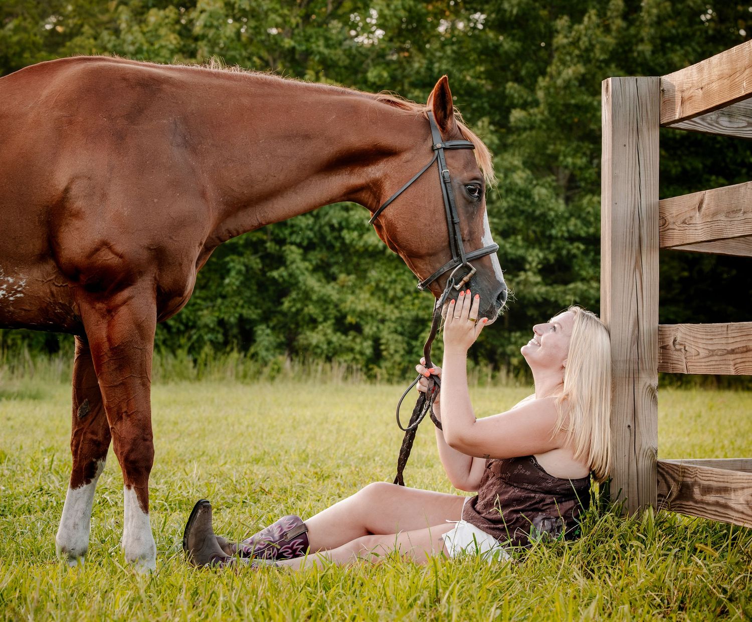 Blonde woman sitting on grass leaning against wooden fence, affectionately petting a chestnut horse wearing a halter.