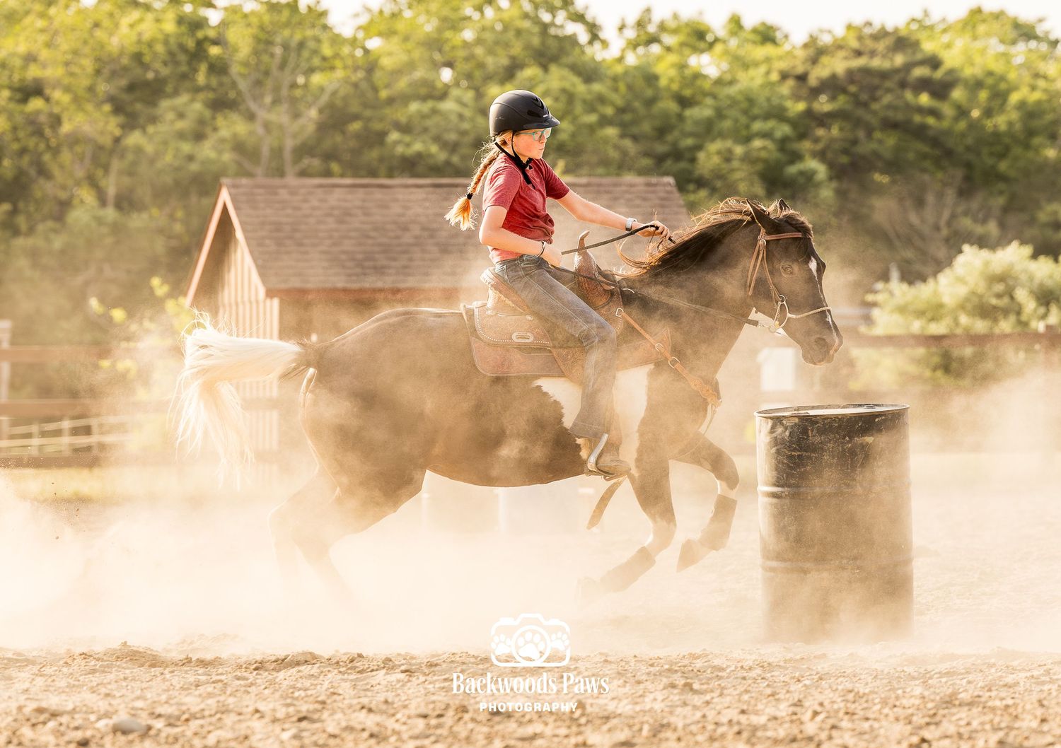 Young rider in red shirt navigates a brown horse around a barrel in a dusty outdoor arena.