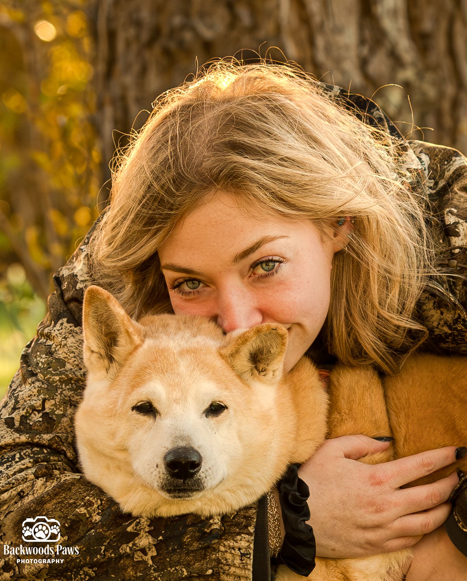 Woman with blonde hair and green eyes hugging a fluffy Shiba Inu dog outdoors in golden autumn light.