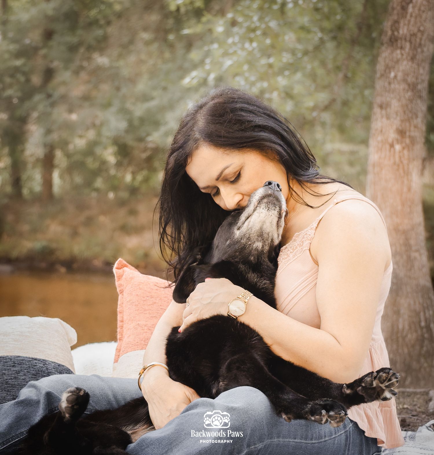 Woman tenderly hugging her large black dog outdoors among trees, captured by Backwoods Paws Photography.