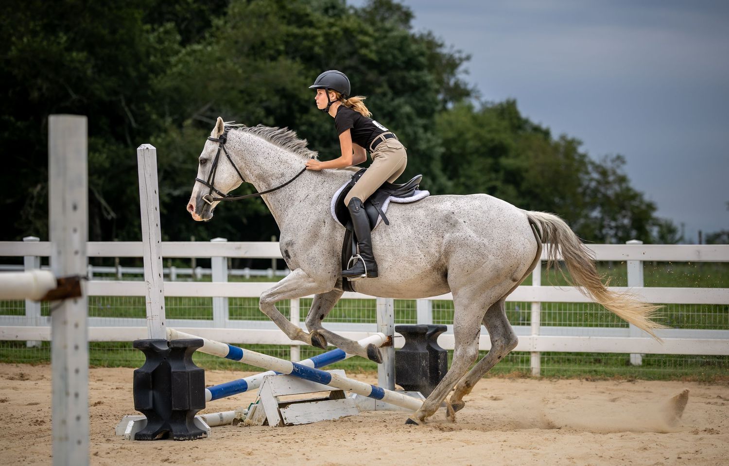 Young equestrian rider jumping a white horse over a colorful hurdle in an outdoor riding arena.