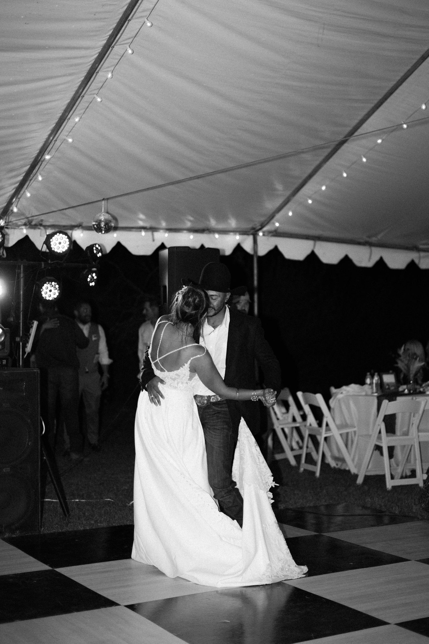 Black and white photo of bride and groom sharing first dance under a tent with string lights at their wedding reception.