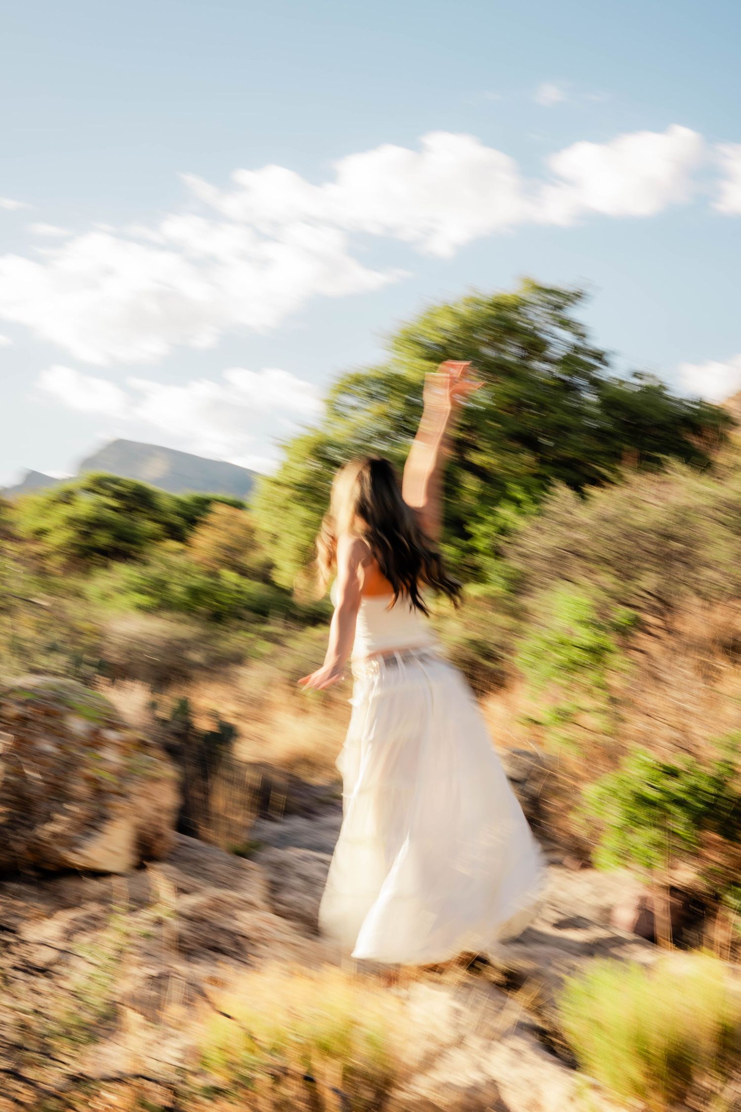 A woman in a white dress dances freely outdoors among rocky terrain and green trees under a bright blue sky.