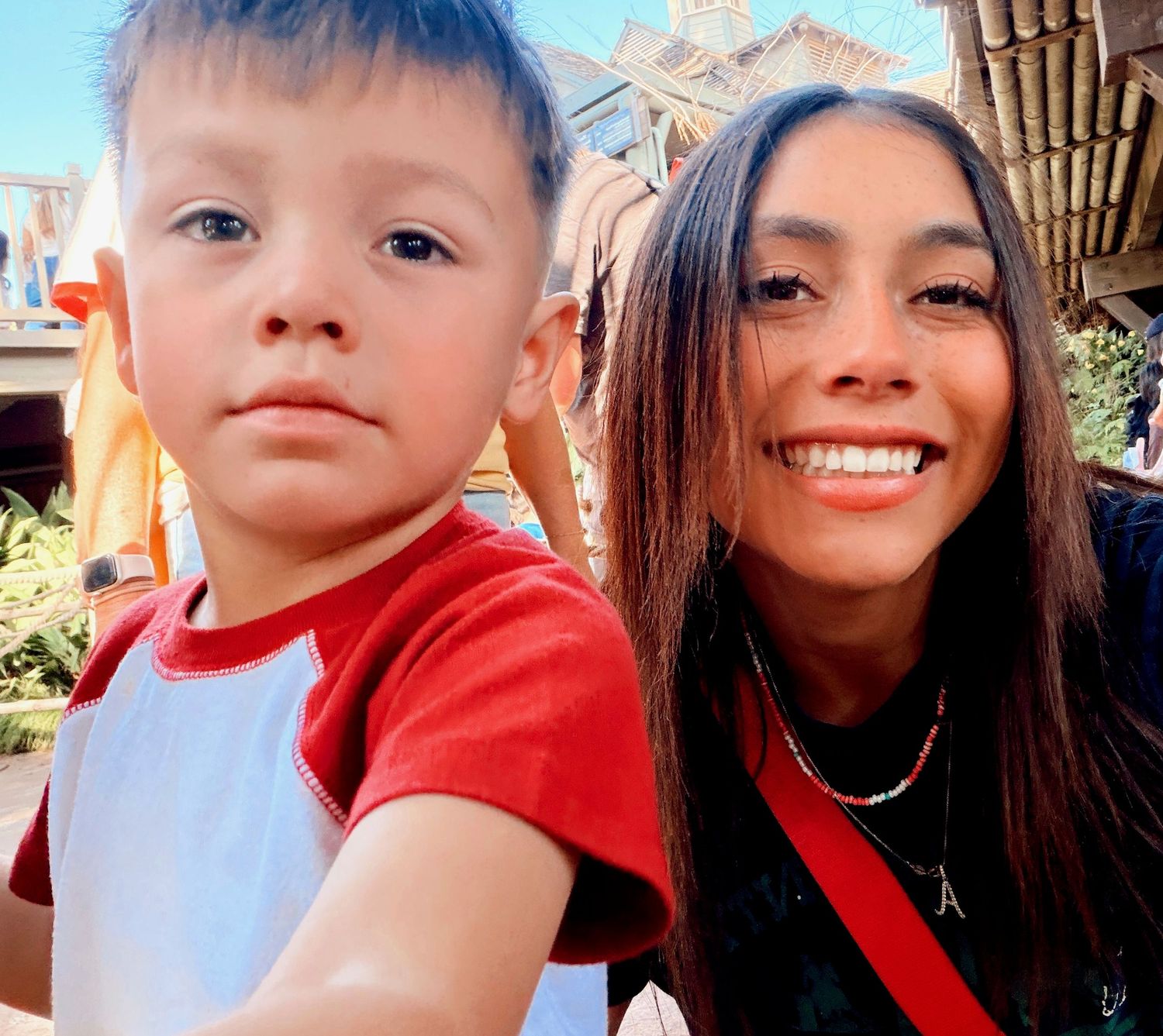 Smiling young woman taking a selfie with a young boy in a red shirt at a theme park.