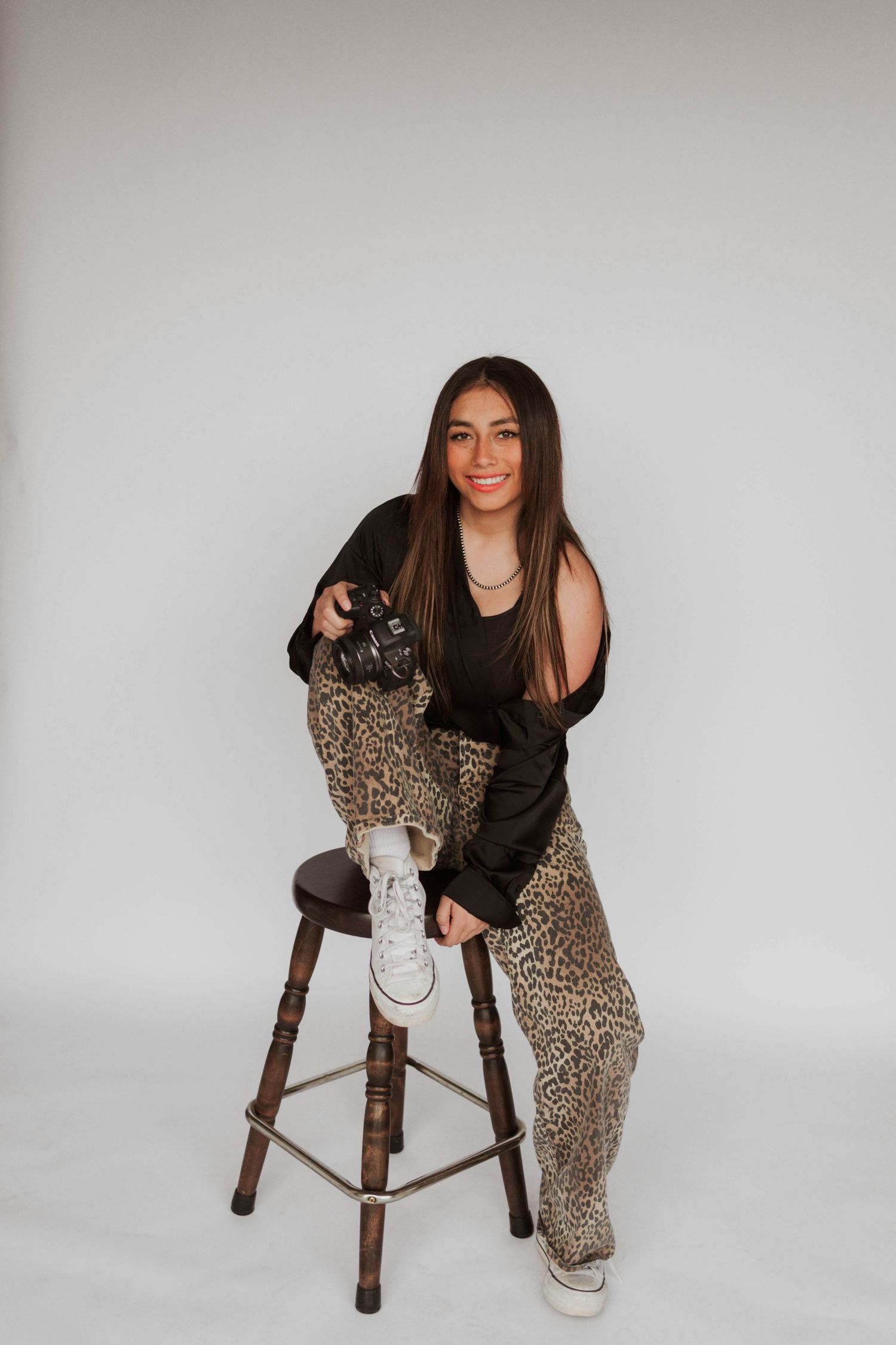 Smiling young woman in leopard print pants holding a camera while sitting on a wooden stool.