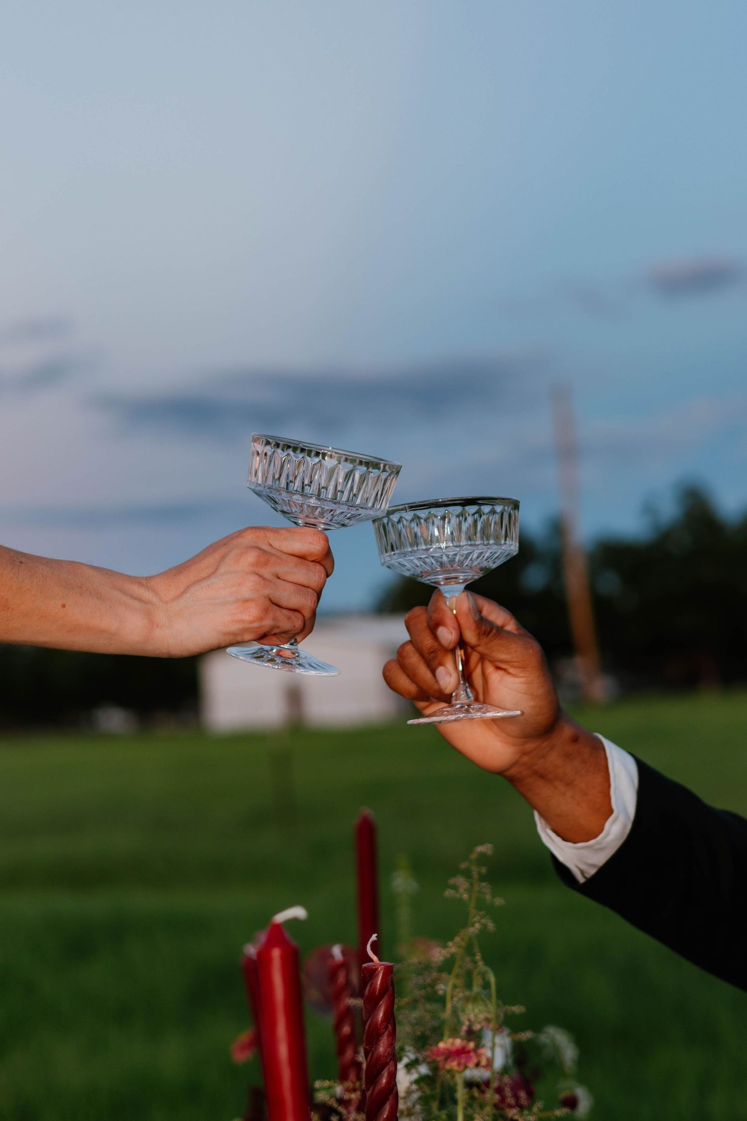 Two hands clinking crystal coupe glasses outdoors at dusk over candlelit table.