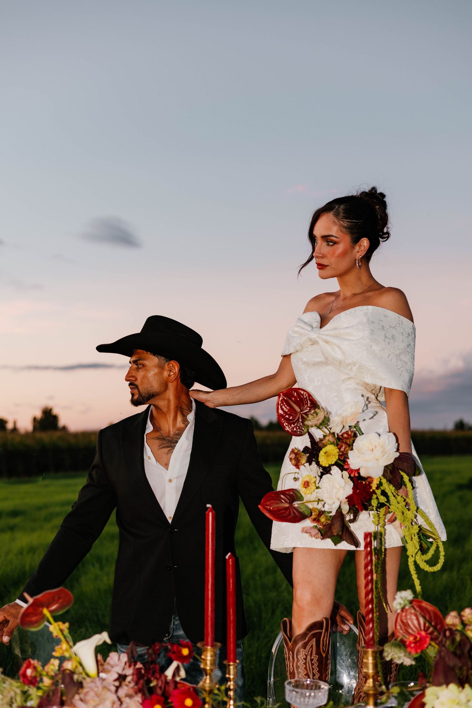 Bride in off-shoulder white mini dress with bouquet stands beside groom in black cowboy hat at sunset.