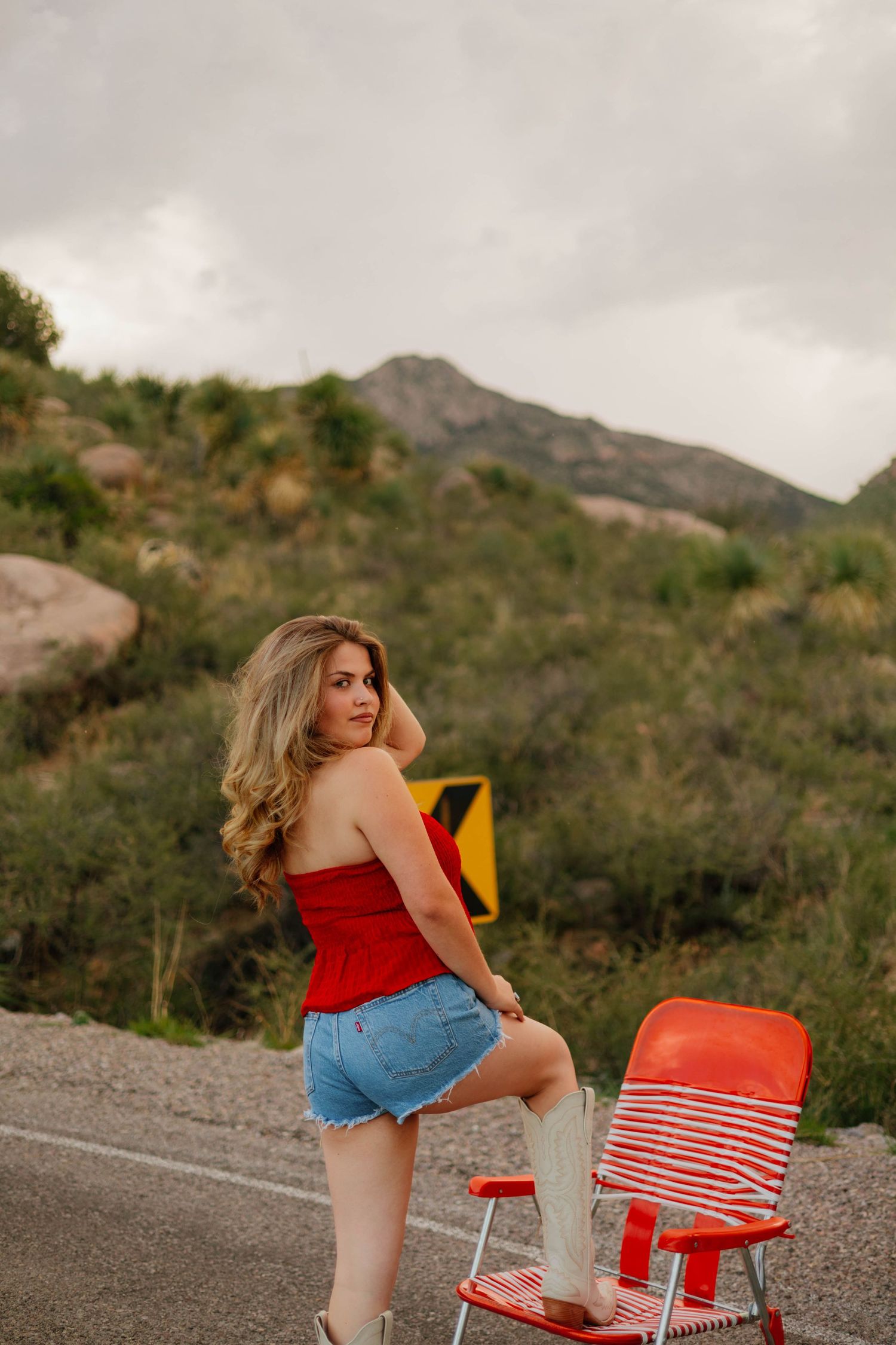Young woman in red tube top and denim shorts poses by a red chair on a mountain road.
