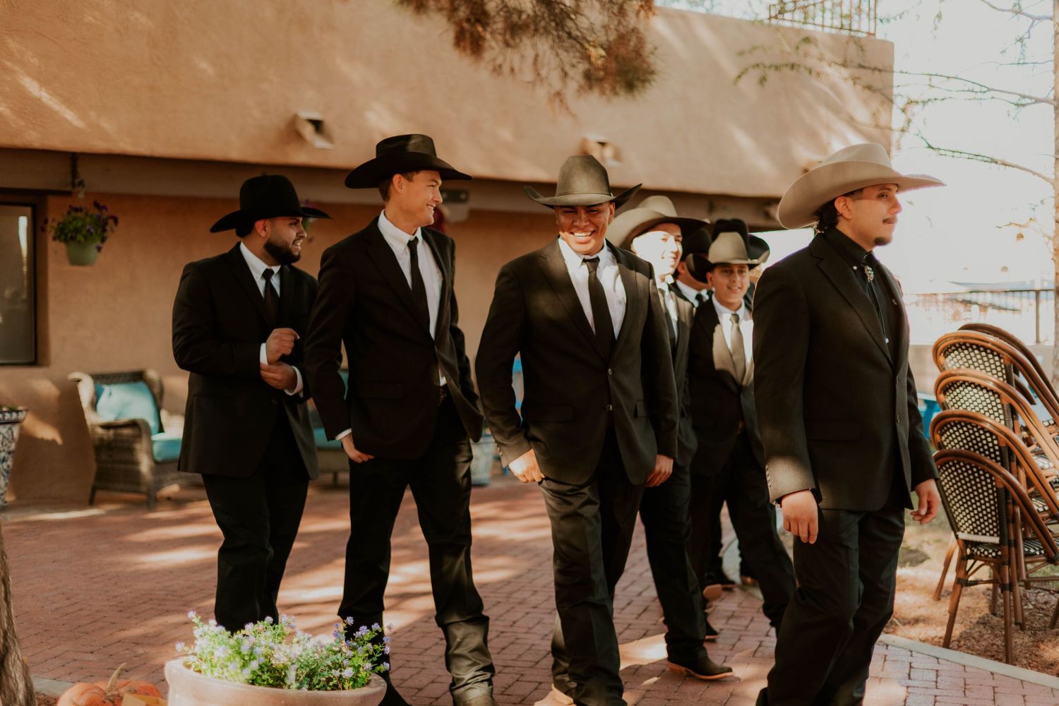 Six men in black suits and cowboy hats walk together outdoors at a rustic western-themed wedding venue.