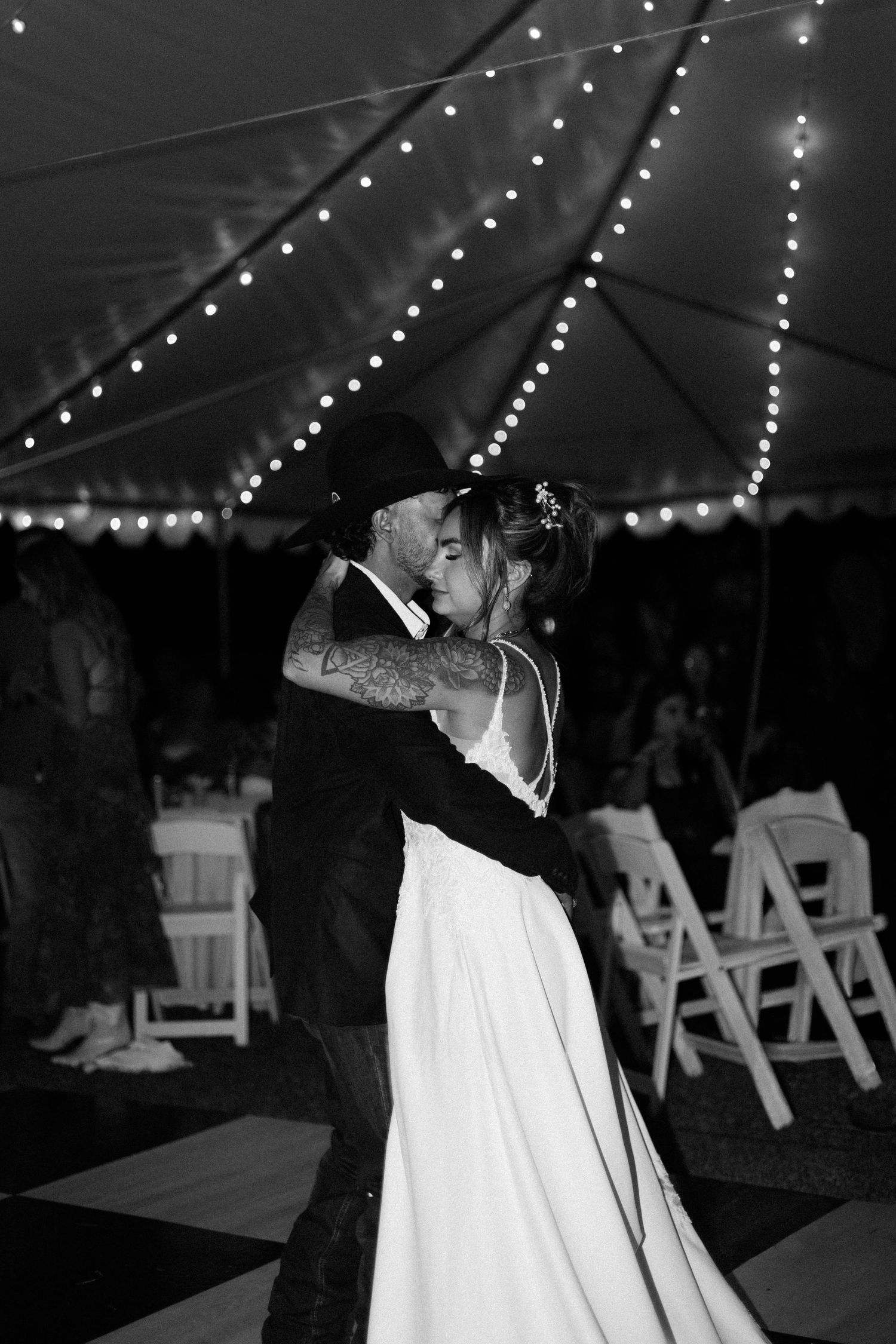 Black and white photo of a bride and groom sharing their first dance under string lights at a tented wedding reception.