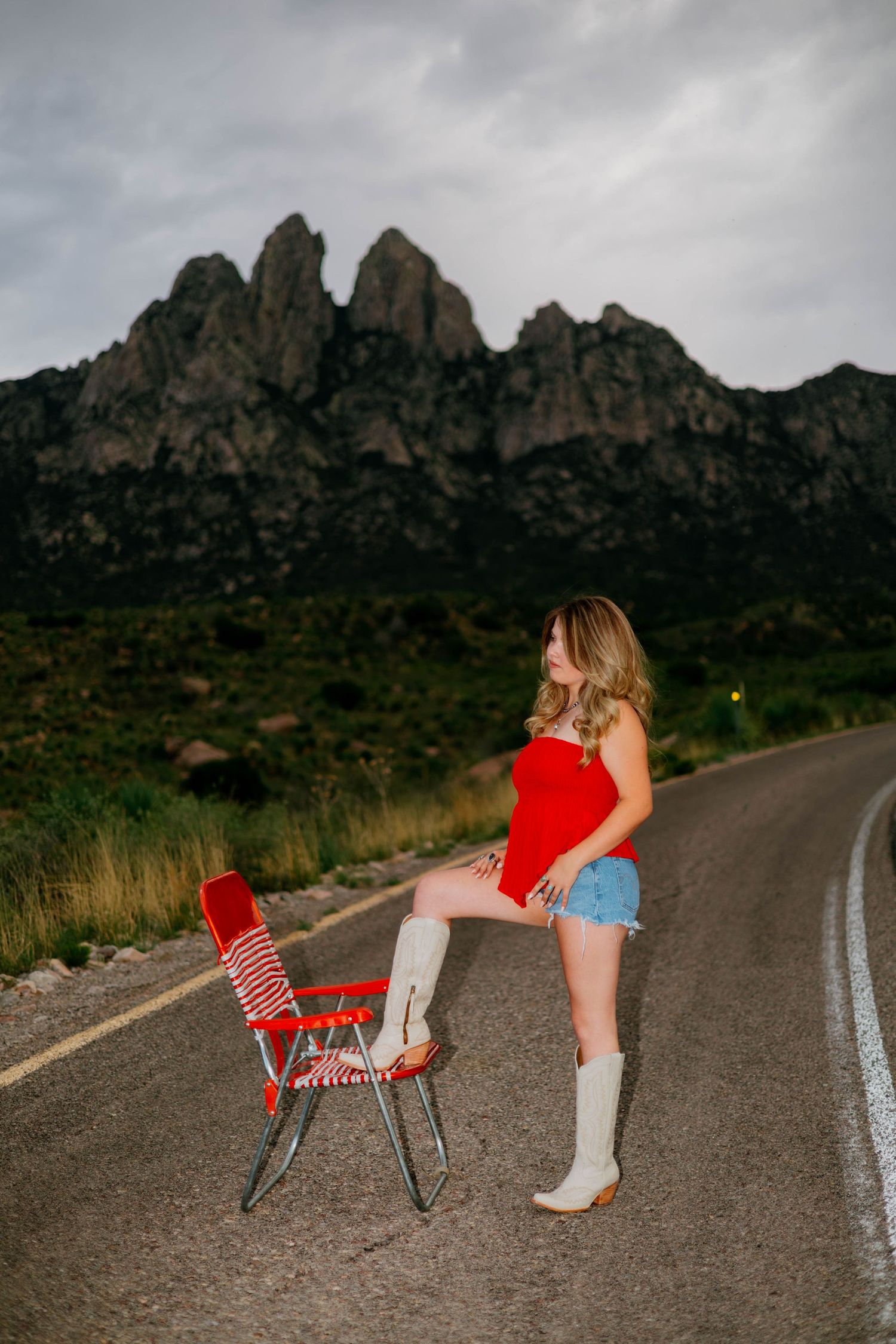 Woman in red top, denim shorts, and white boots poses on a road with dramatic rocky mountains in the background.