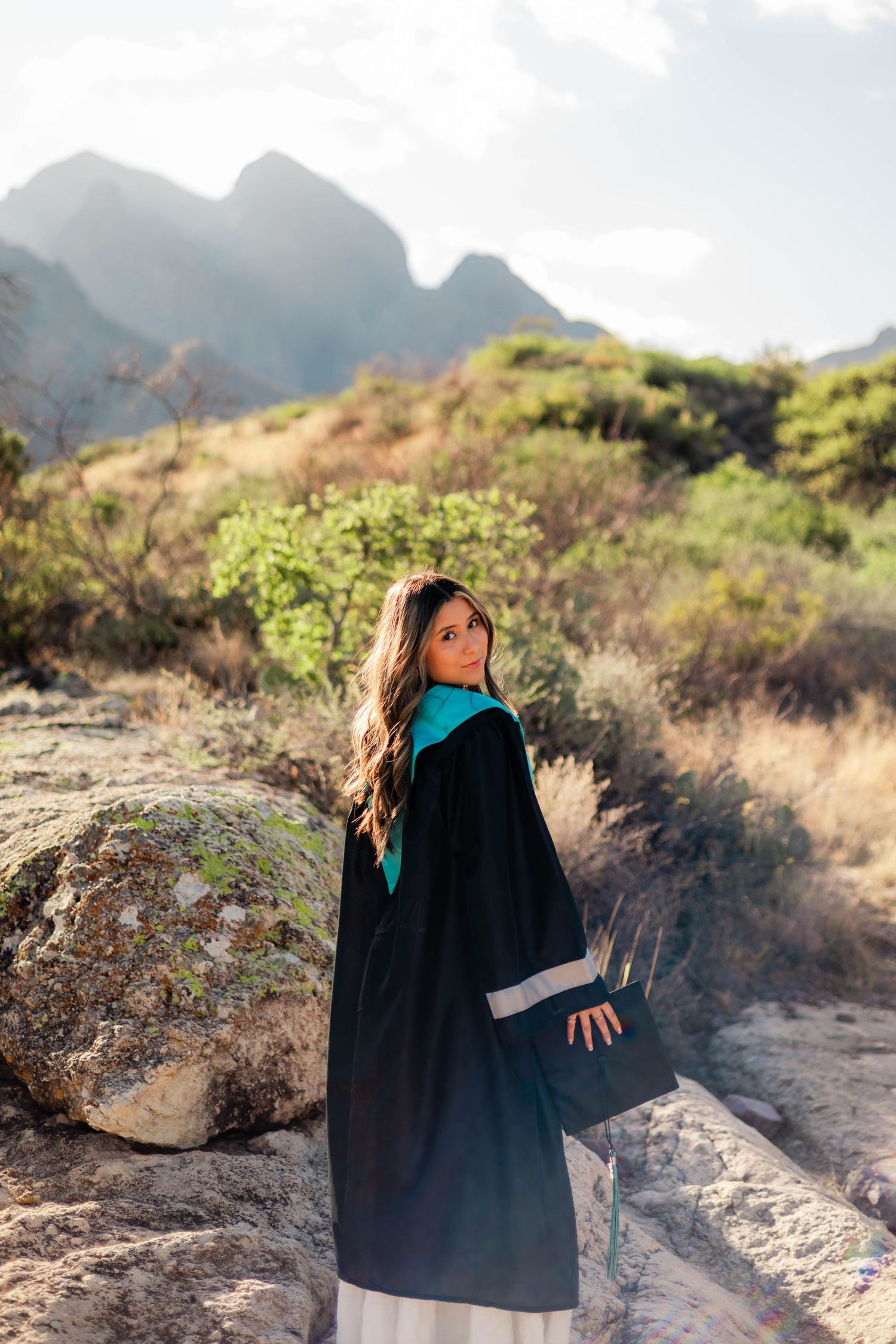Young woman in black graduation gown with teal hood posing on a rocky desert mountain trail.