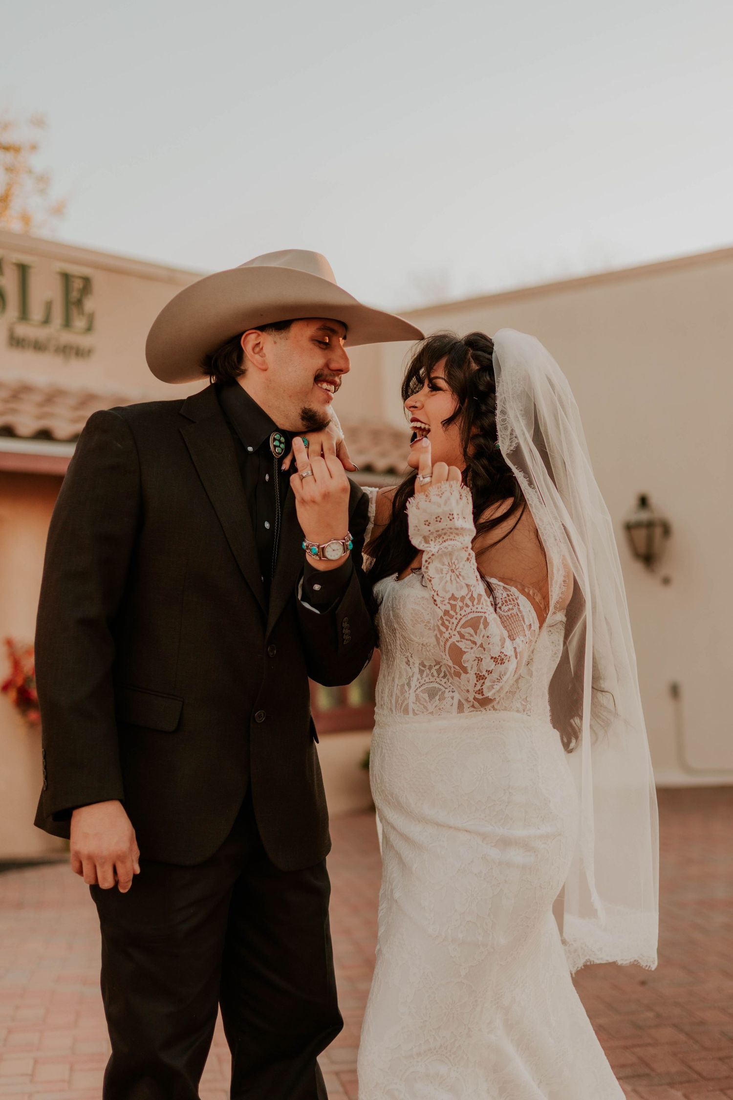 A bride in lace gown and veil stands with groom in black suit and cowboy hat, sharing a playful moment outdoors.