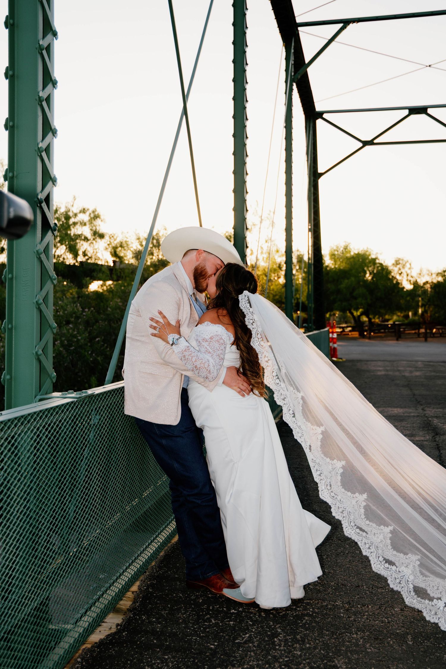 Bride and groom kissing on a green iron bridge with flowing lace veil.