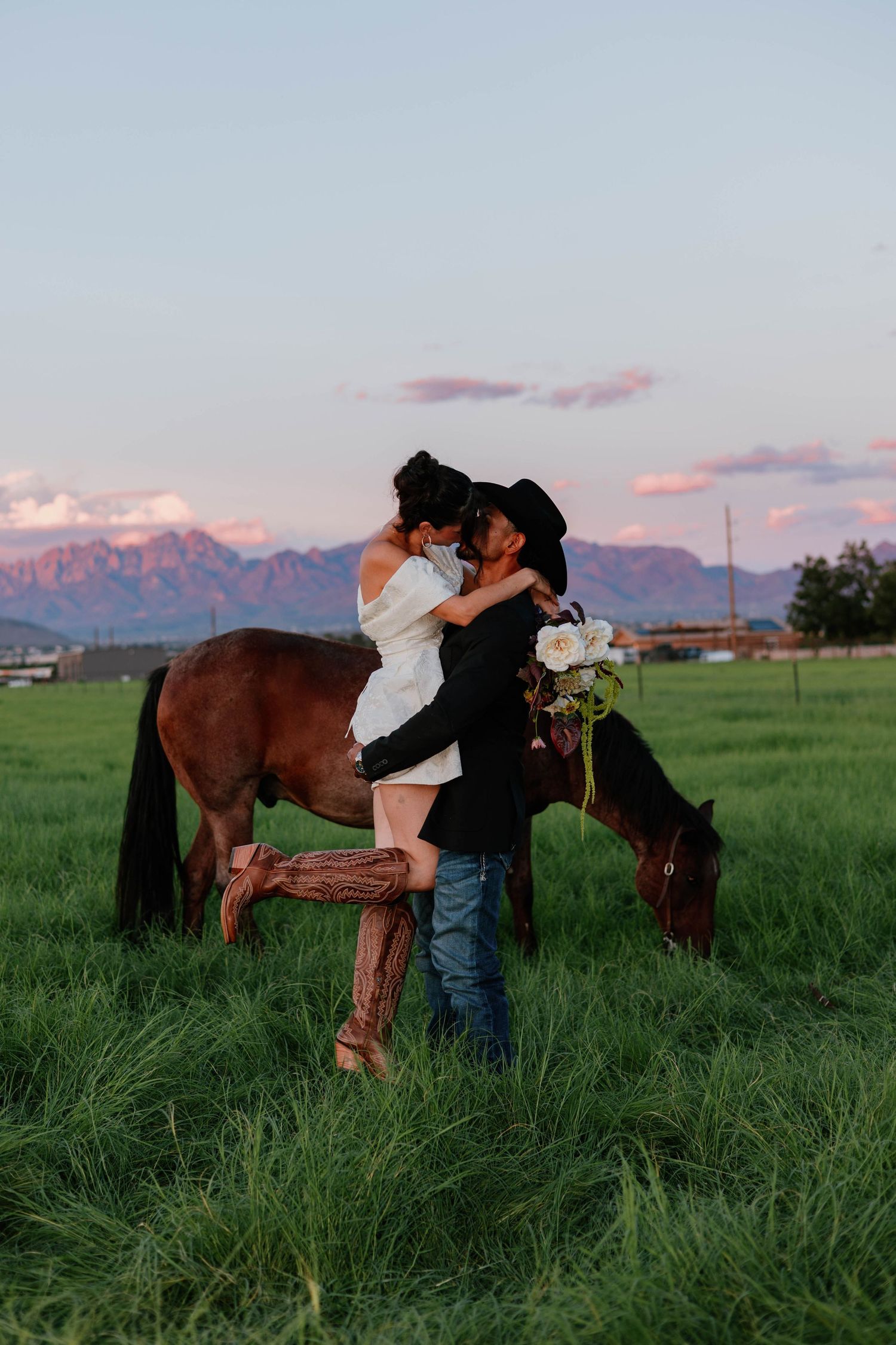 Couple kissing beside a brown horse in a green field at mountain sunset.