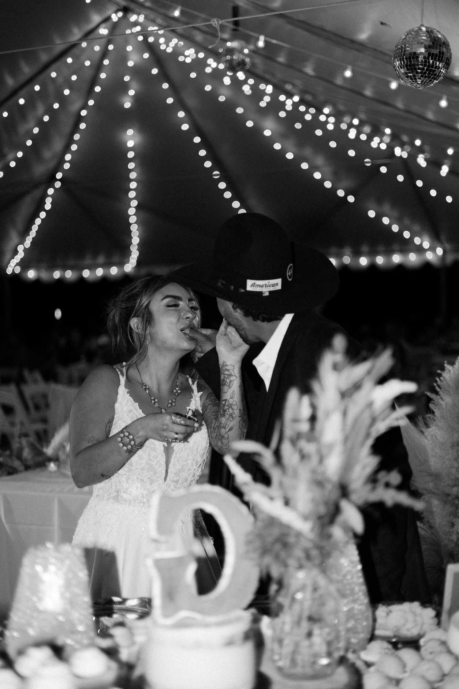 Black and white photo of a couple sharing an intimate moment under fairy lights at a wedding reception.