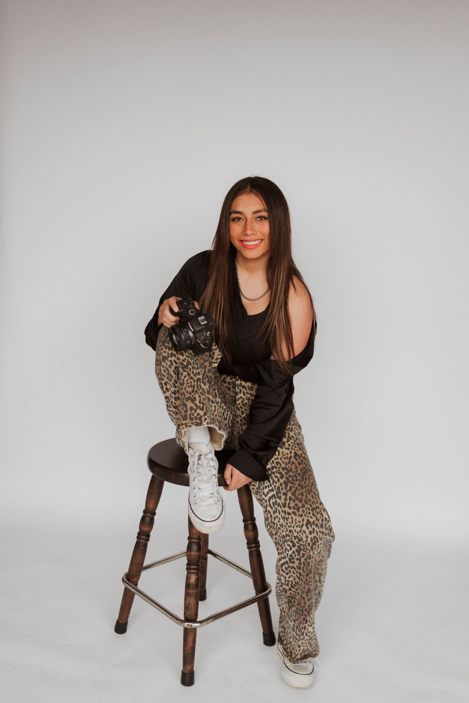 Young woman in leopard print pants and black top sits on wooden stool holding camera in studio.