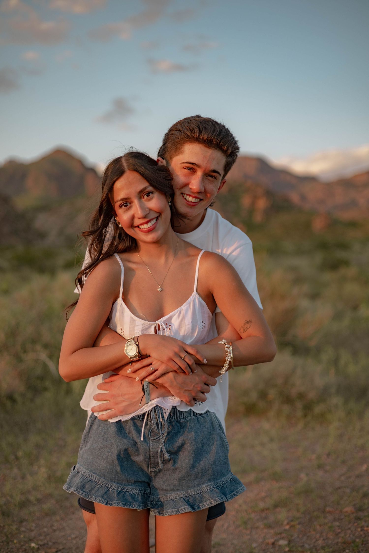 A smiling couple embraces outdoors at golden hour with desert mountains in the background.
