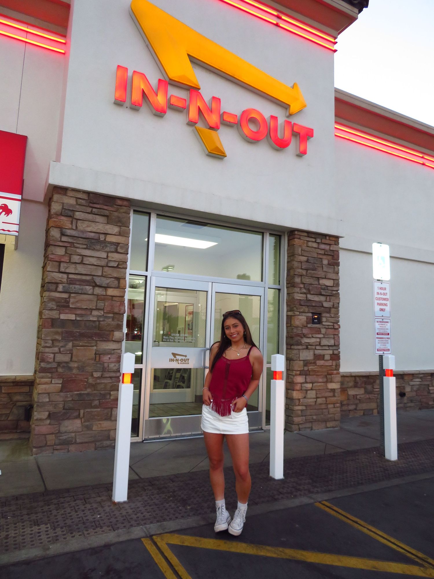 Young woman in red top and white skirt standing outside an In-N-Out Burger restaurant entrance.