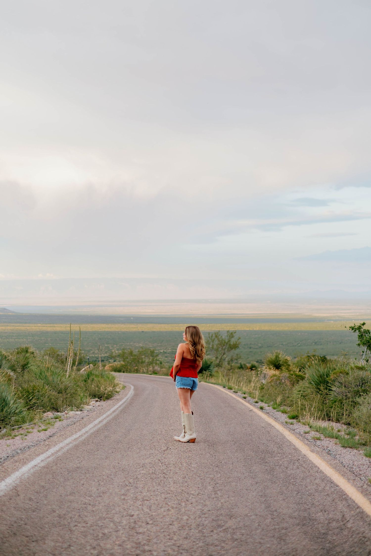 Woman in red top, denim shorts, and white cowboy boots walks alone on a scenic desert highway.