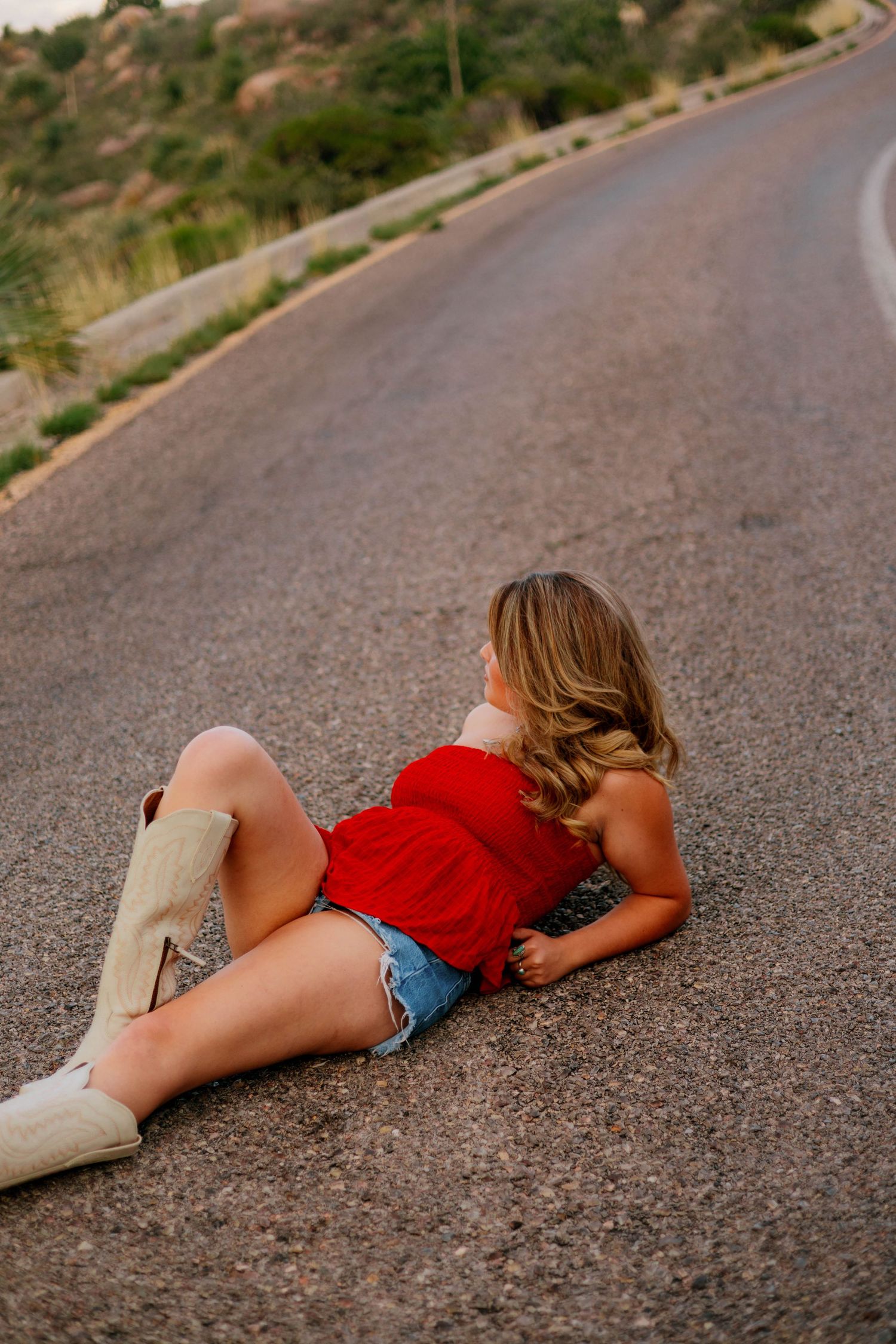 Young woman in red top, denim shorts, and cowboy boots lying on a rural road, looking away from camera.