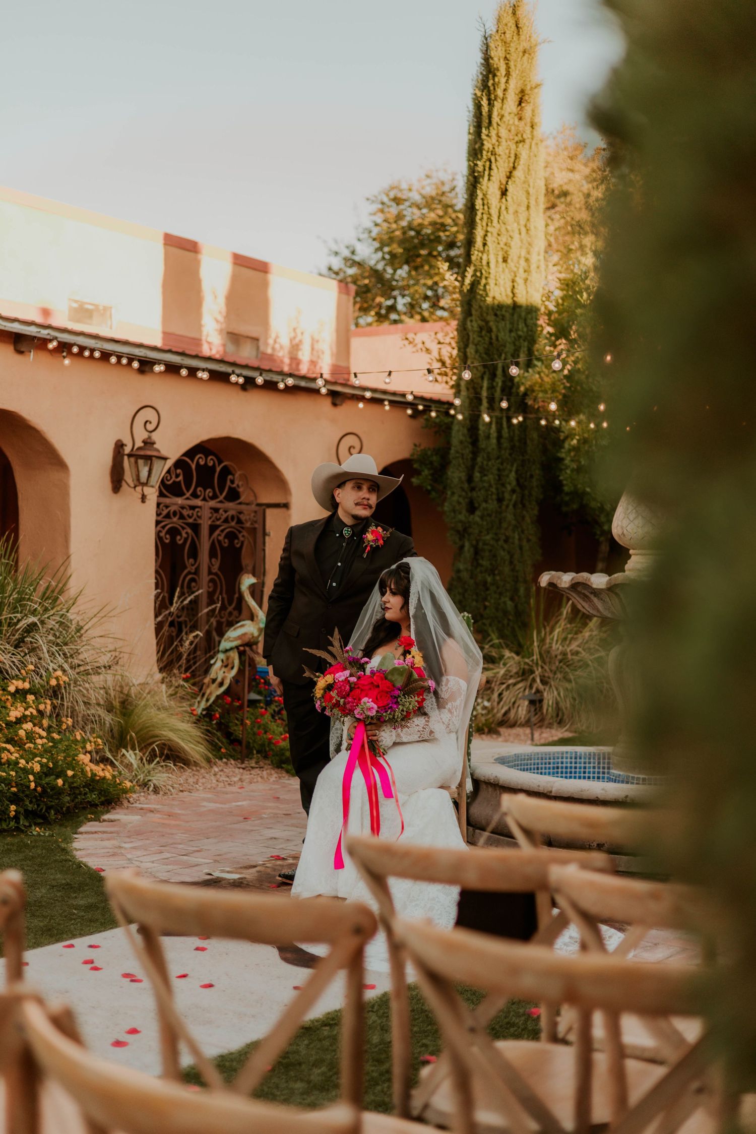 Bride in white gown with pink bouquet walks down outdoor aisle with groom in black suit and cowboy hat at sunset wedding.