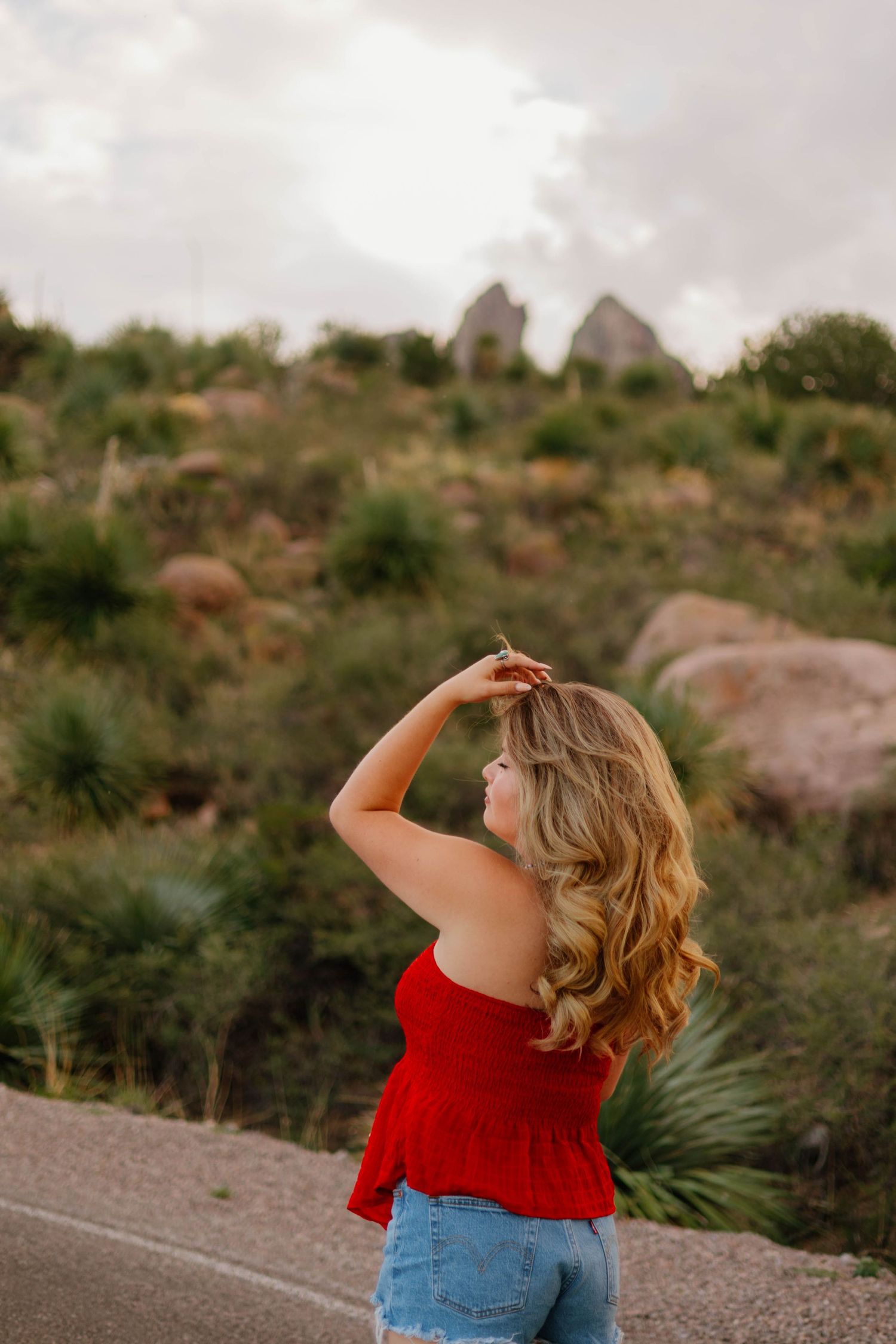 Blonde woman in red top and denim shorts poses outdoors near rocky desert landscape with lush greenery.