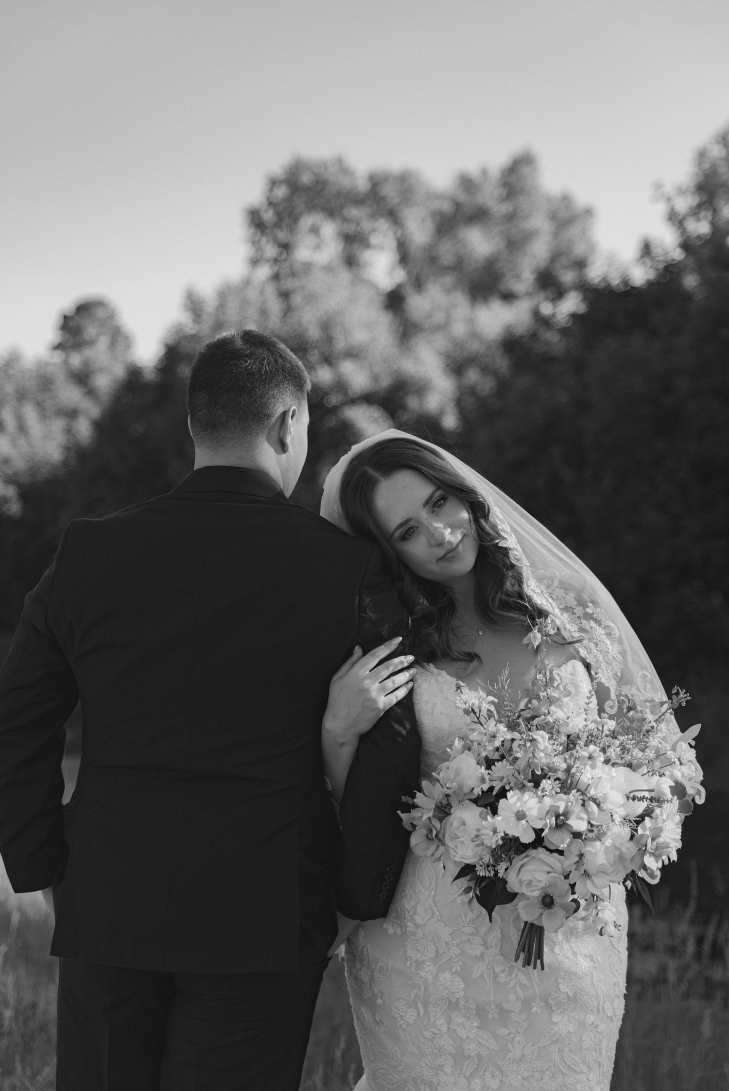 Black and white photo of a bride holding a bouquet, leaning into her groom outdoors on their wedding day.