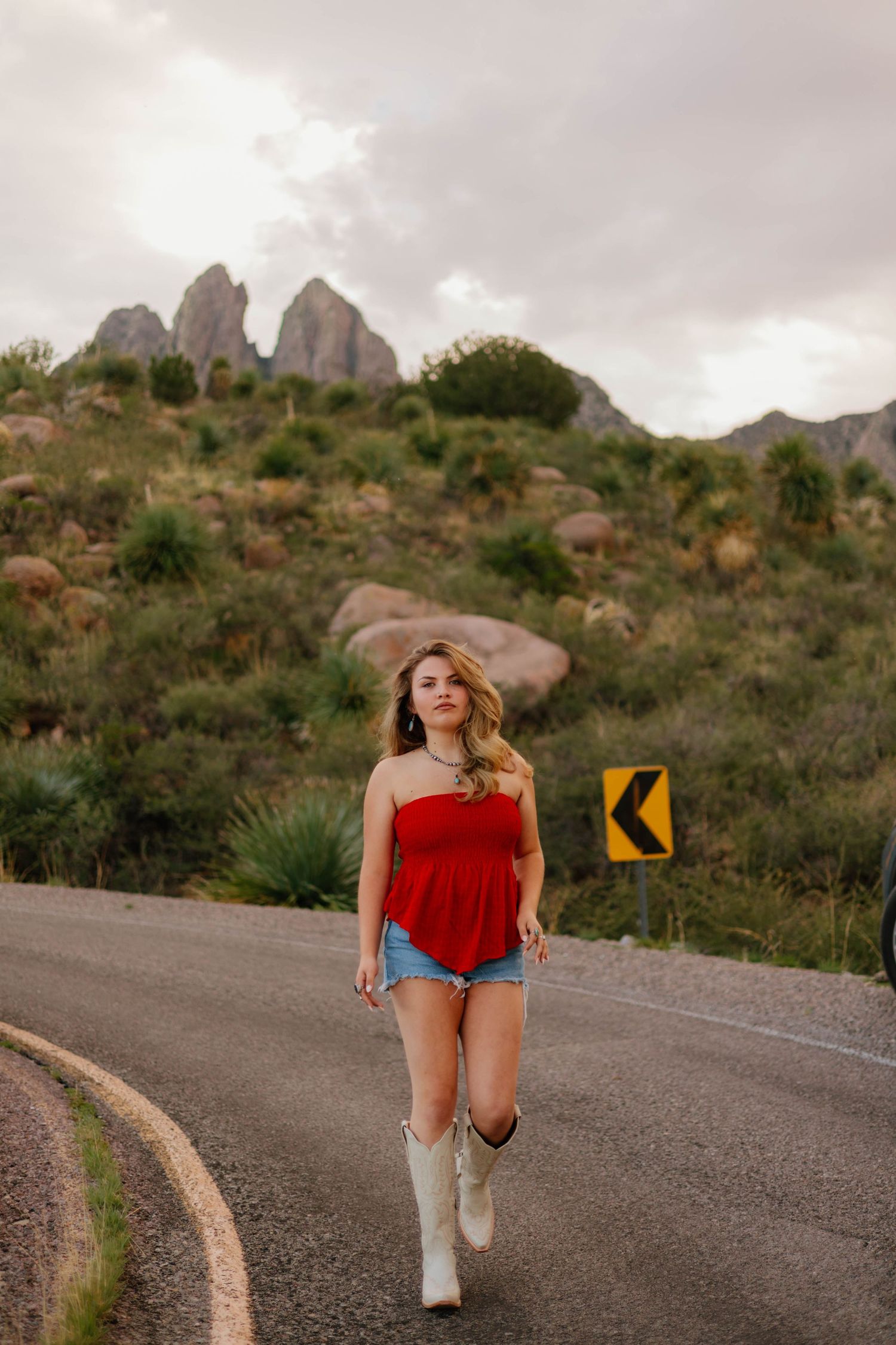 Young woman in red top, denim shorts, and white cowboy boots walking on a scenic mountain road.