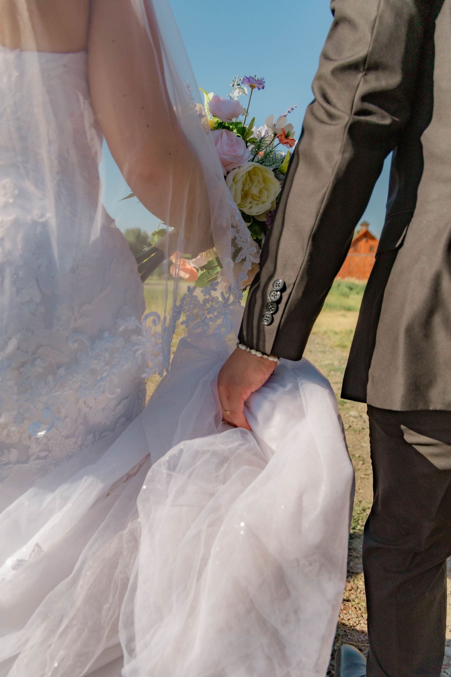Bride and groom holding hands, bride in white lace gown with veil holding floral bouquet outdoors on sunny wedding day.