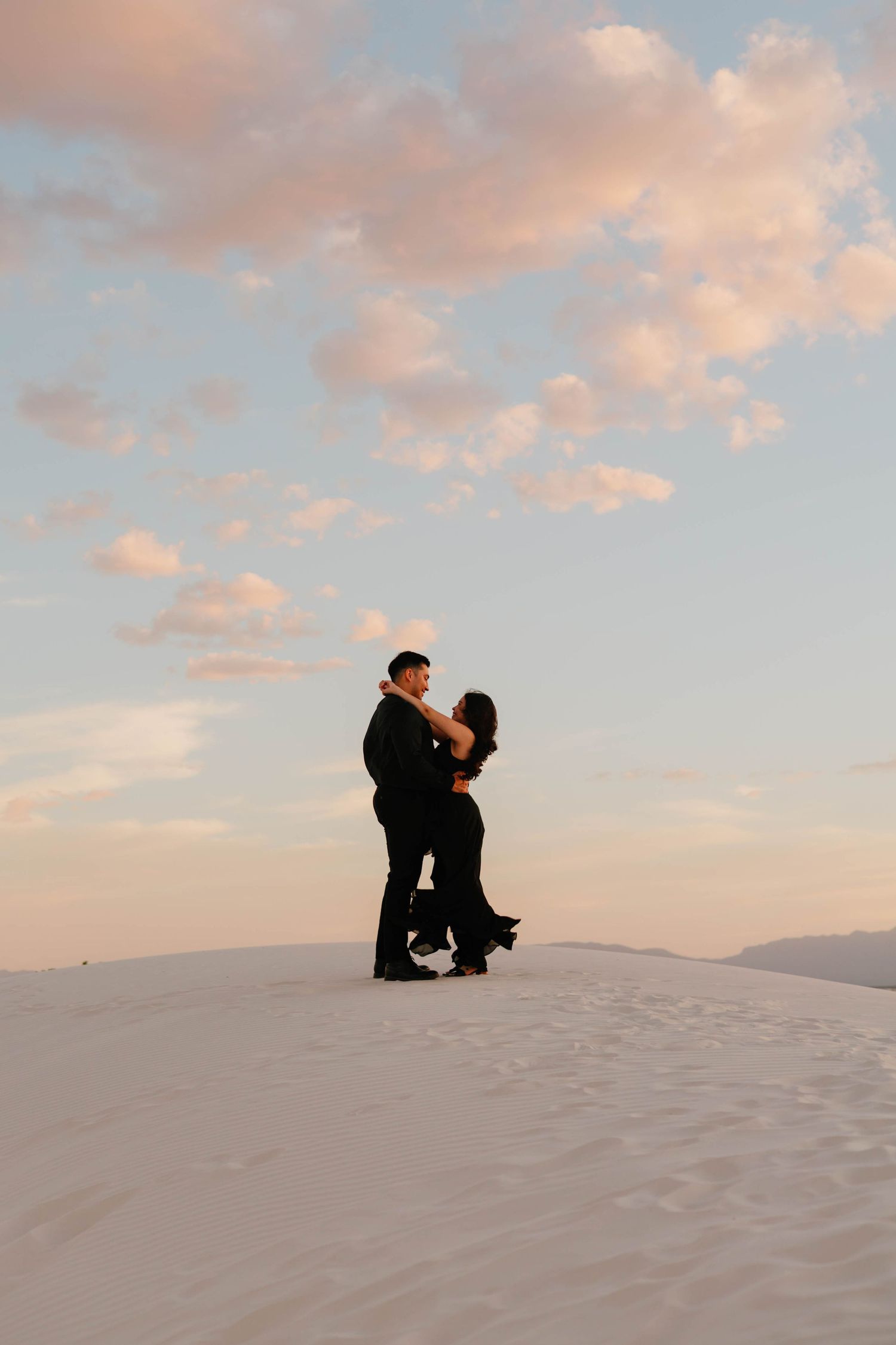 Couple dancing romantically on white sand dunes silhouetted against a pastel sunset sky.