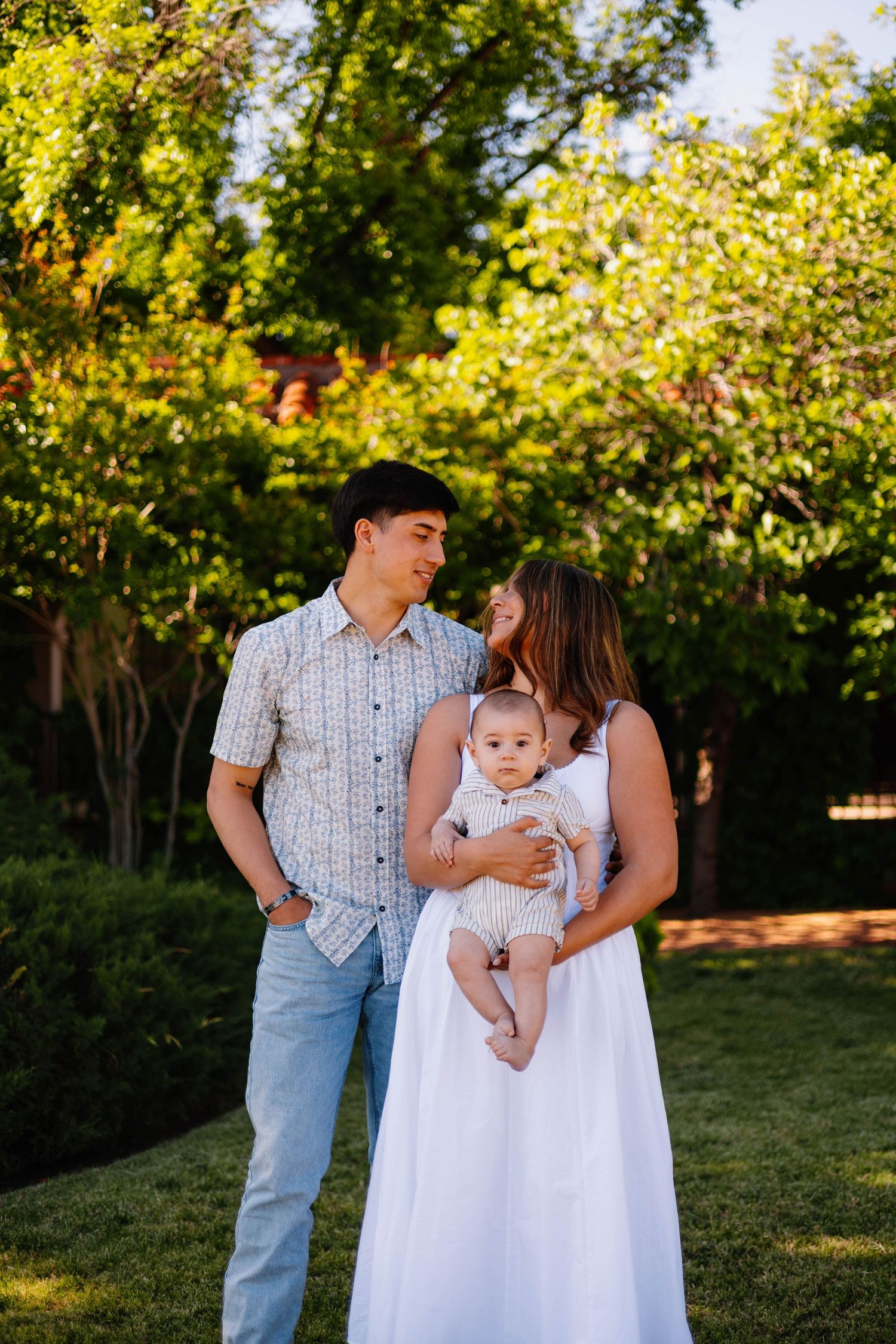 Young family with a baby posing outdoors in a lush green garden during golden hour.