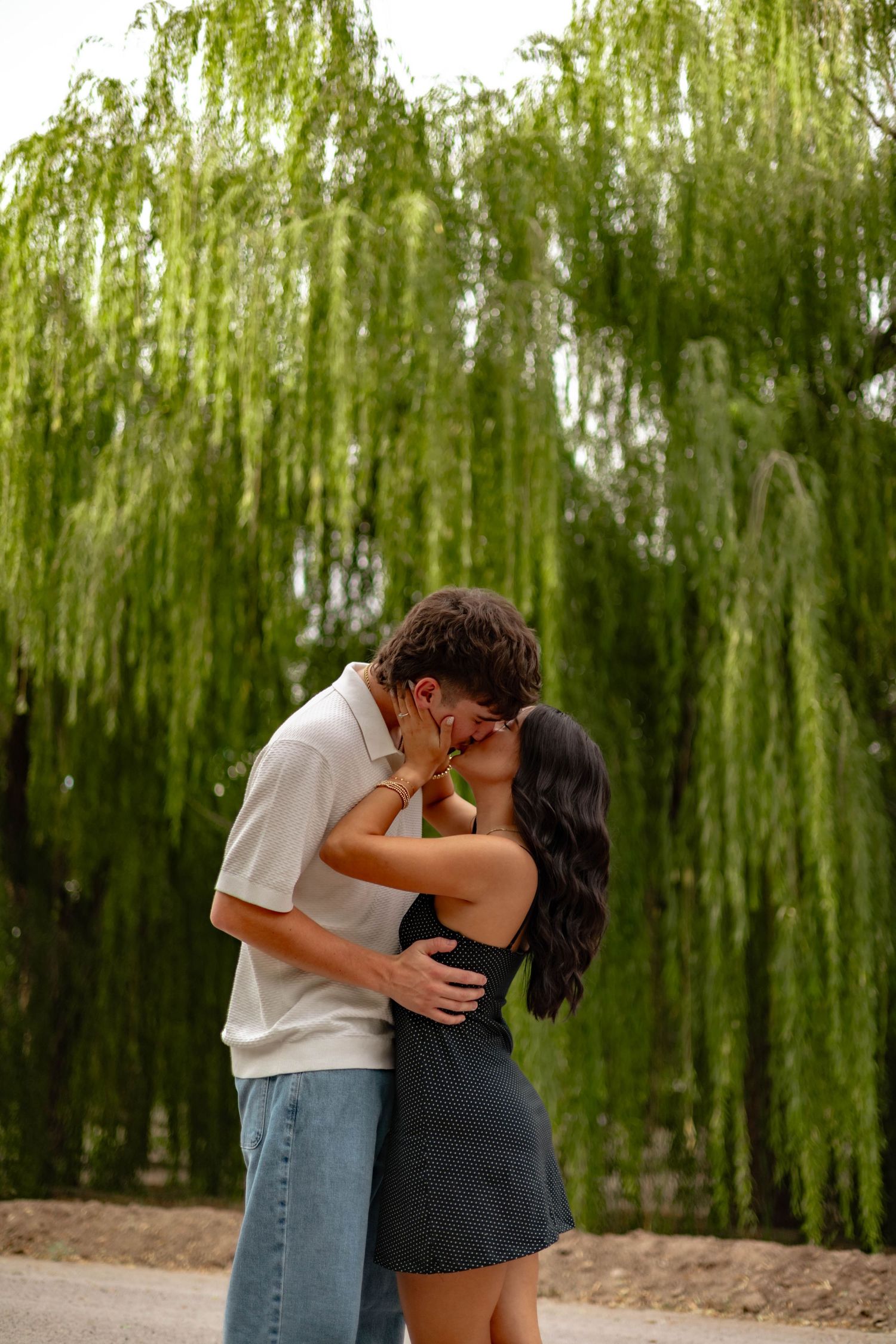 A couple shares a romantic kiss outdoors beneath a lush weeping willow tree, with the woman in a black mini dress.