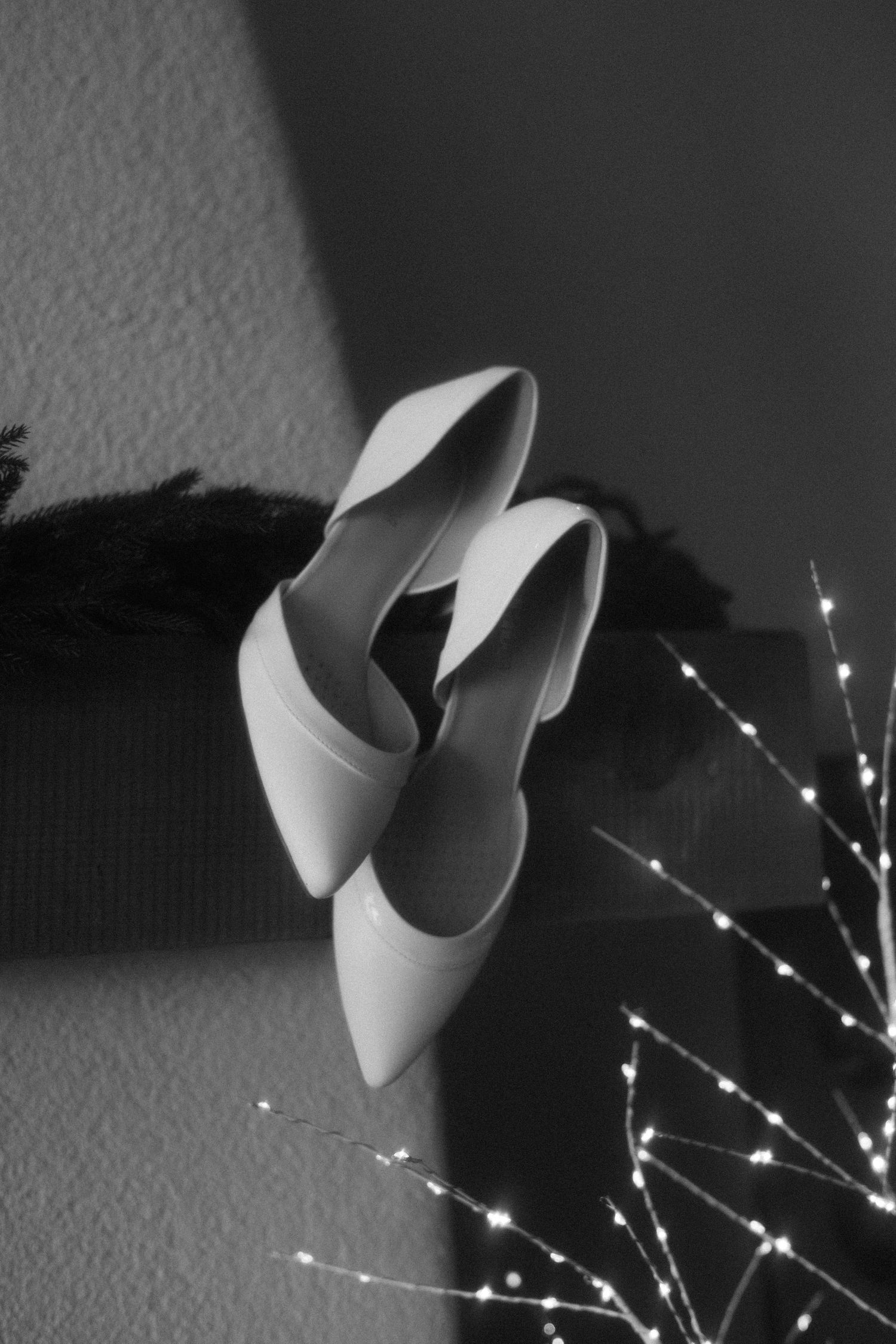 Black and white photo of elegant white pointed-toe heels resting near fairy lights and pine branches.