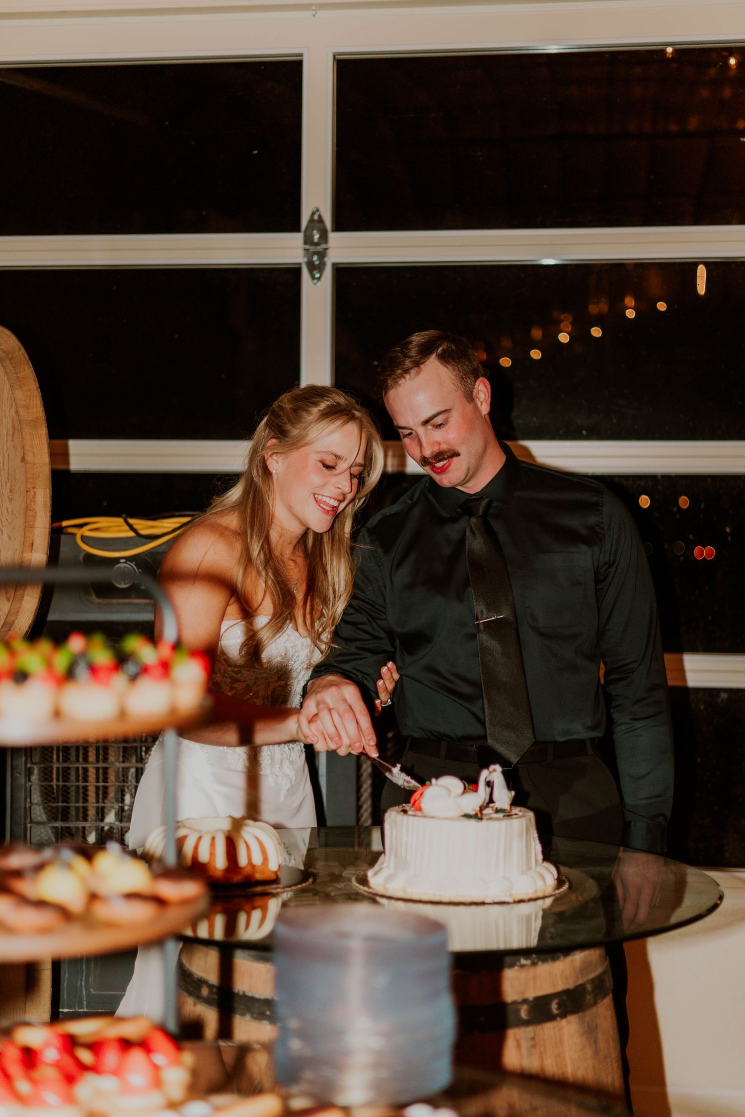 Bride and groom smiling while cutting their white wedding cake at a romantic evening reception.