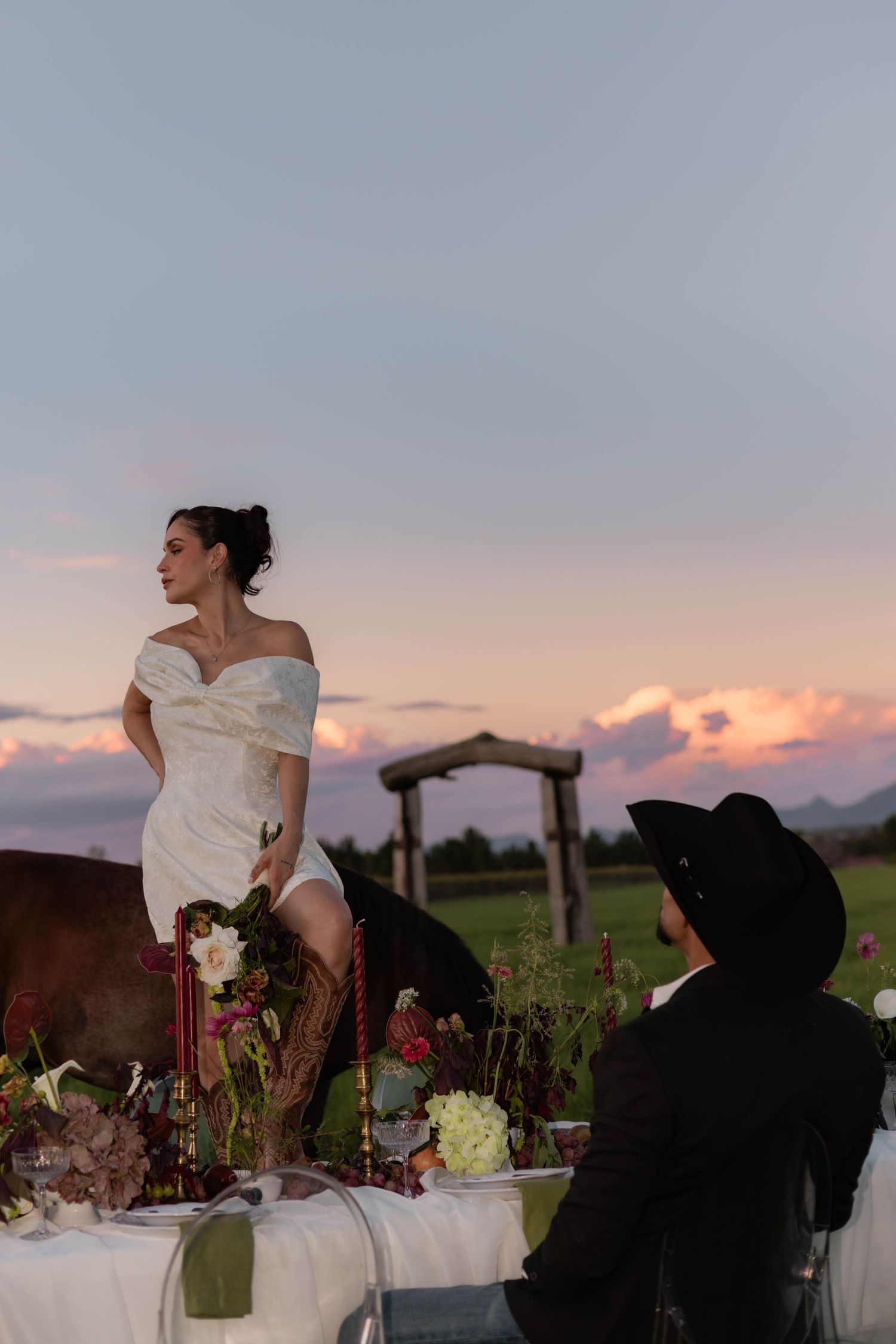 Bride in white off-shoulder dress stands on table at outdoor wedding reception at sunset, cowboy guest watches.