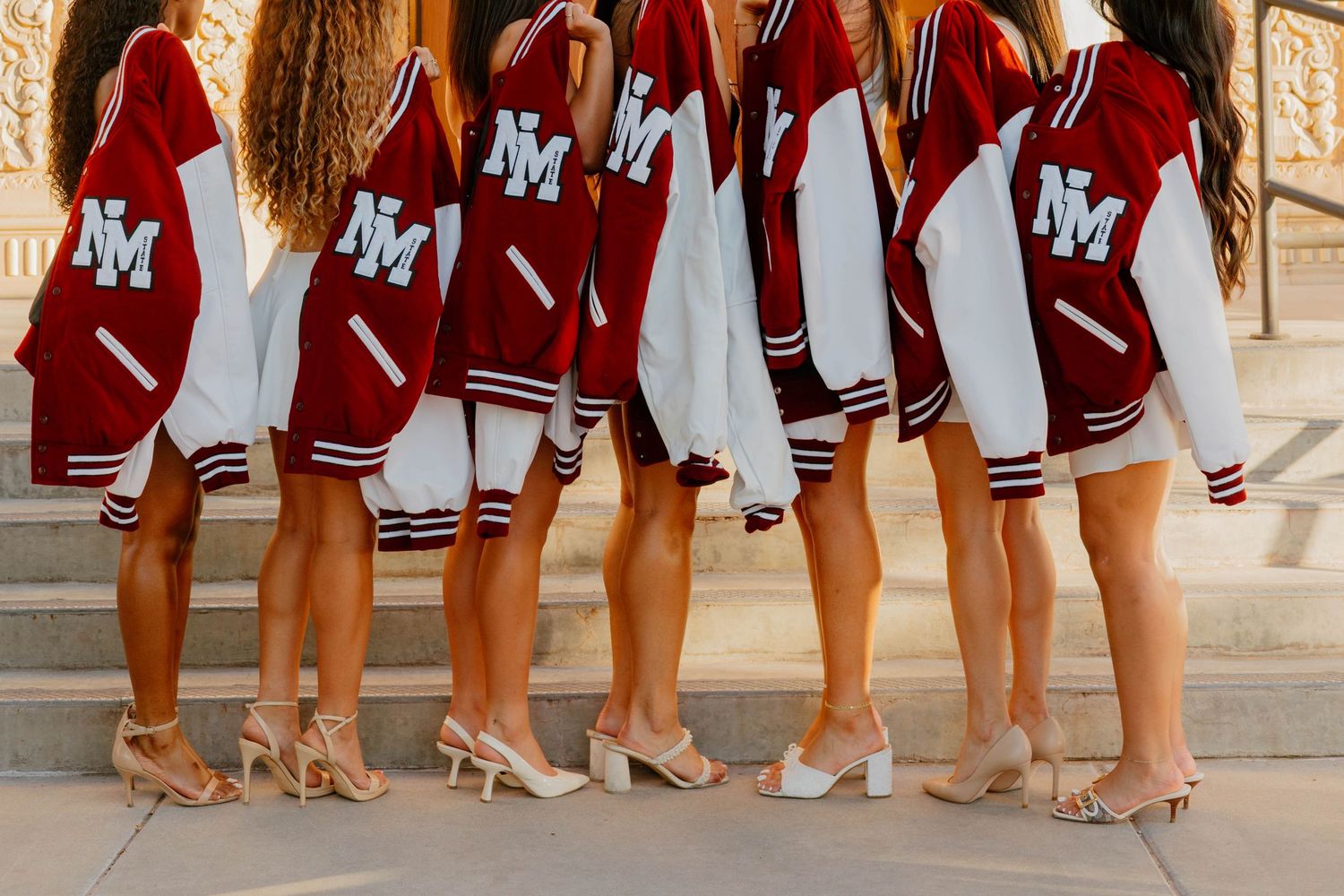 Six women posing in matching red and white NM varsity letterman jackets with heels on steps.