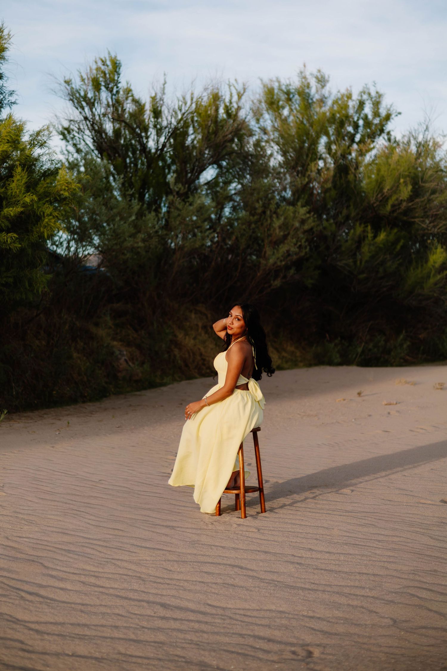 Woman in yellow dress seated on stool in sandy desert landscape at sunset.