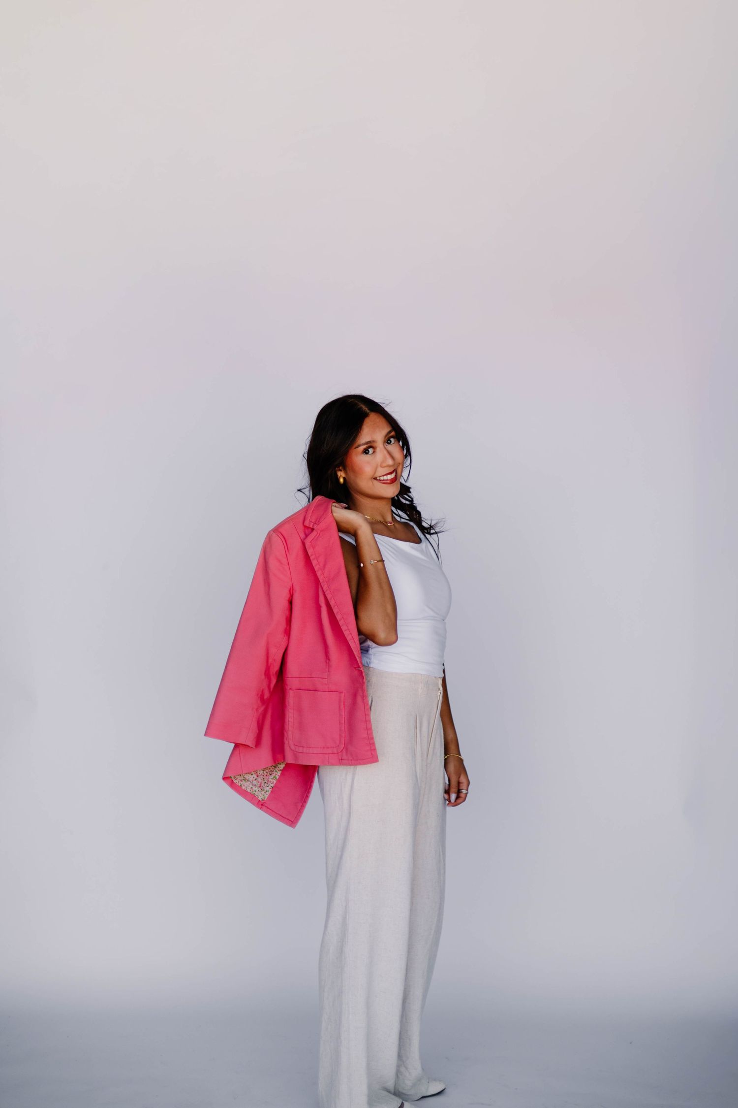 Smiling woman in white outfit holding pink blazer over shoulder against clean white studio background.