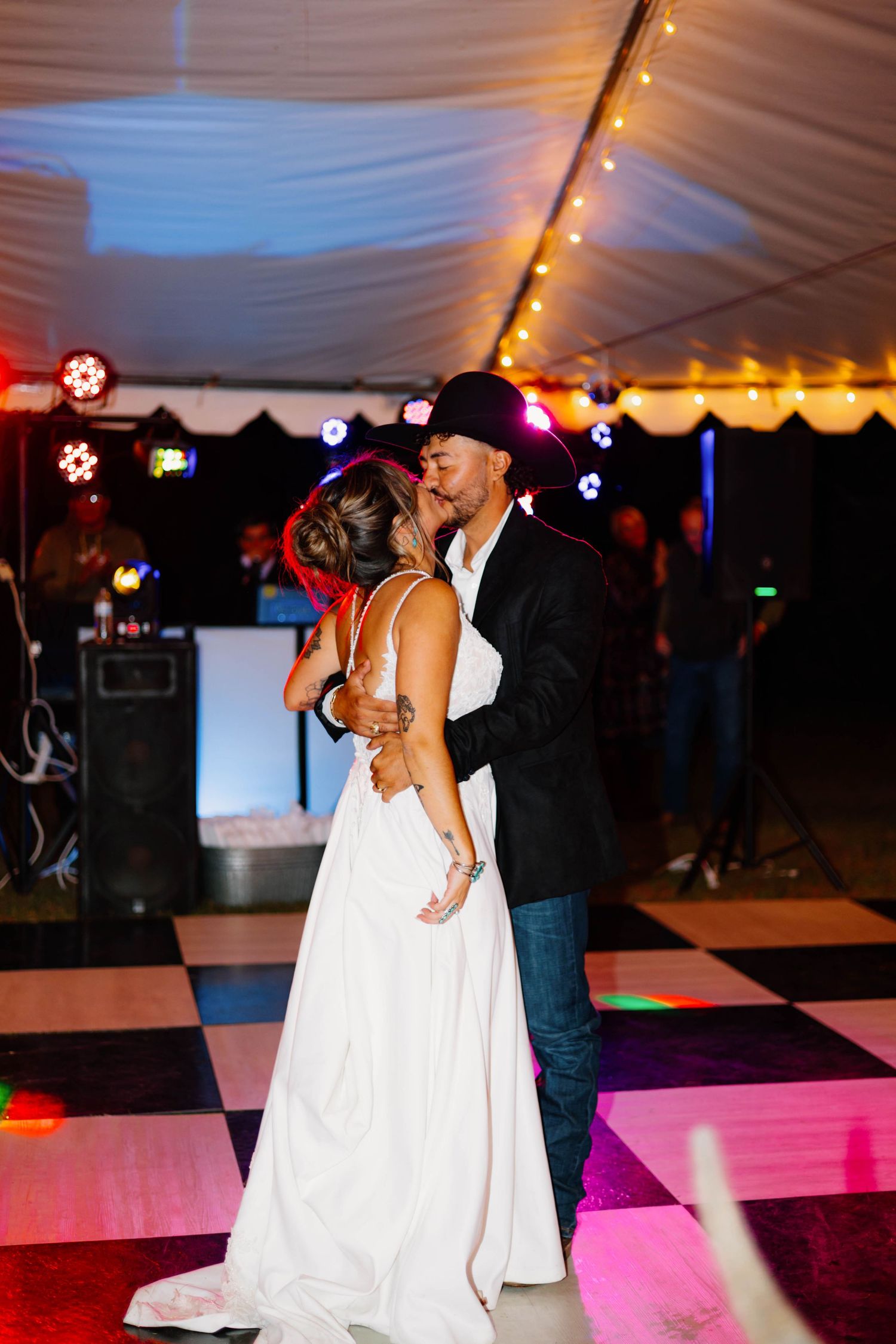 Bride and groom share first dance kiss on black and white checkered floor under tent with colorful wedding reception lights.