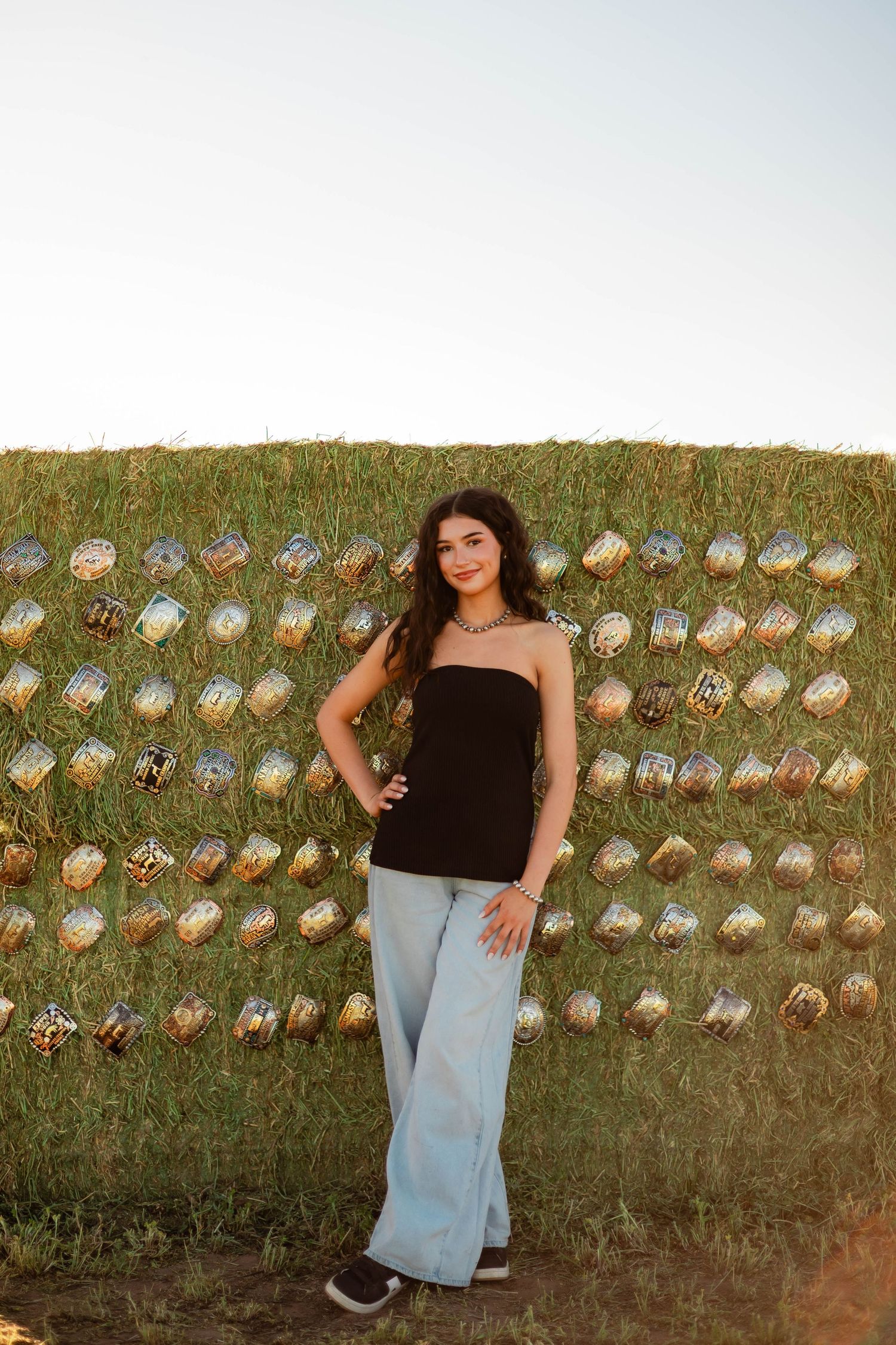 Young woman in black top and wide-leg jeans posing before a hay bale wall.