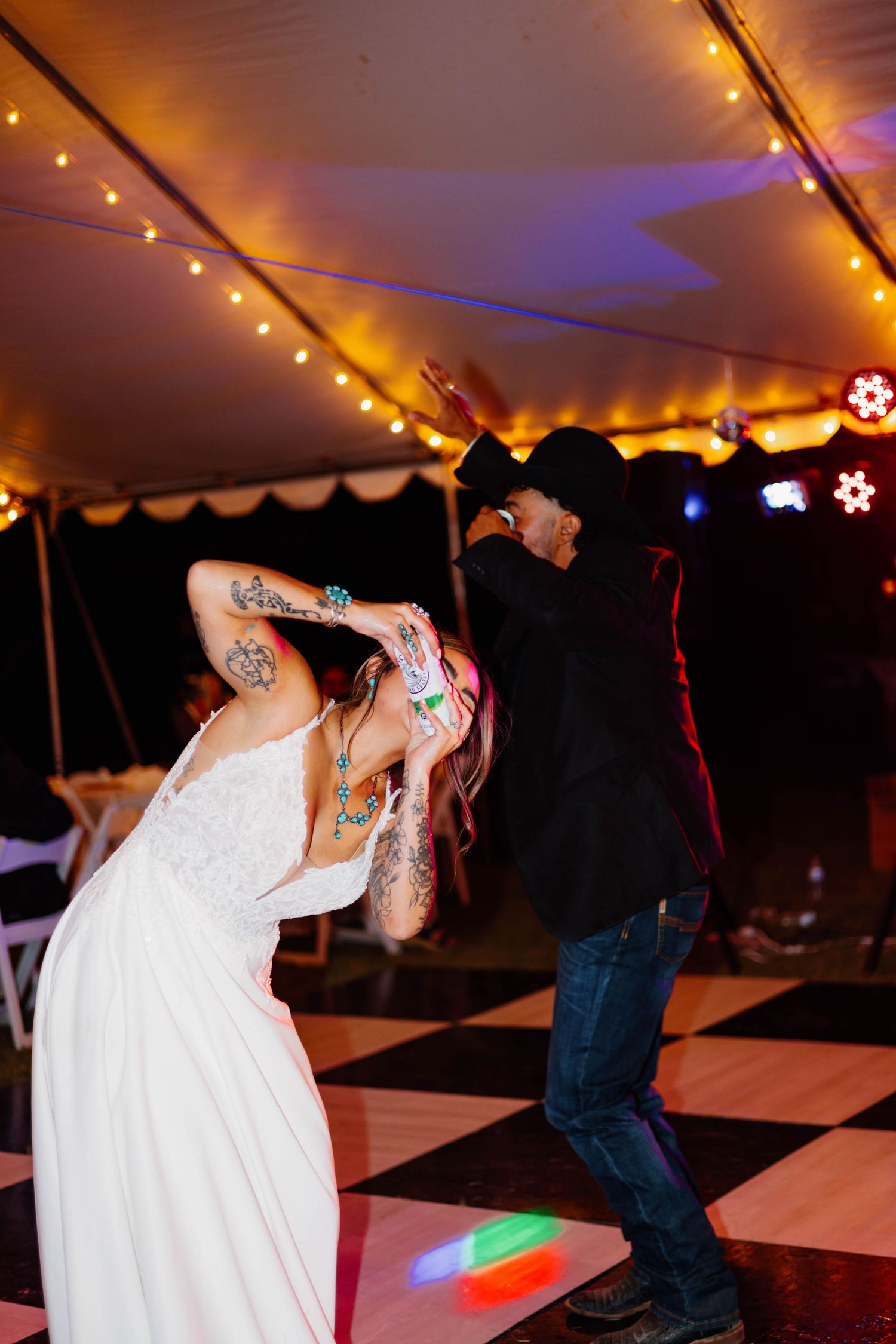 Tattooed bride in white dress dancing with groom in black jacket on checkered dance floor under string lights at wedding reception.